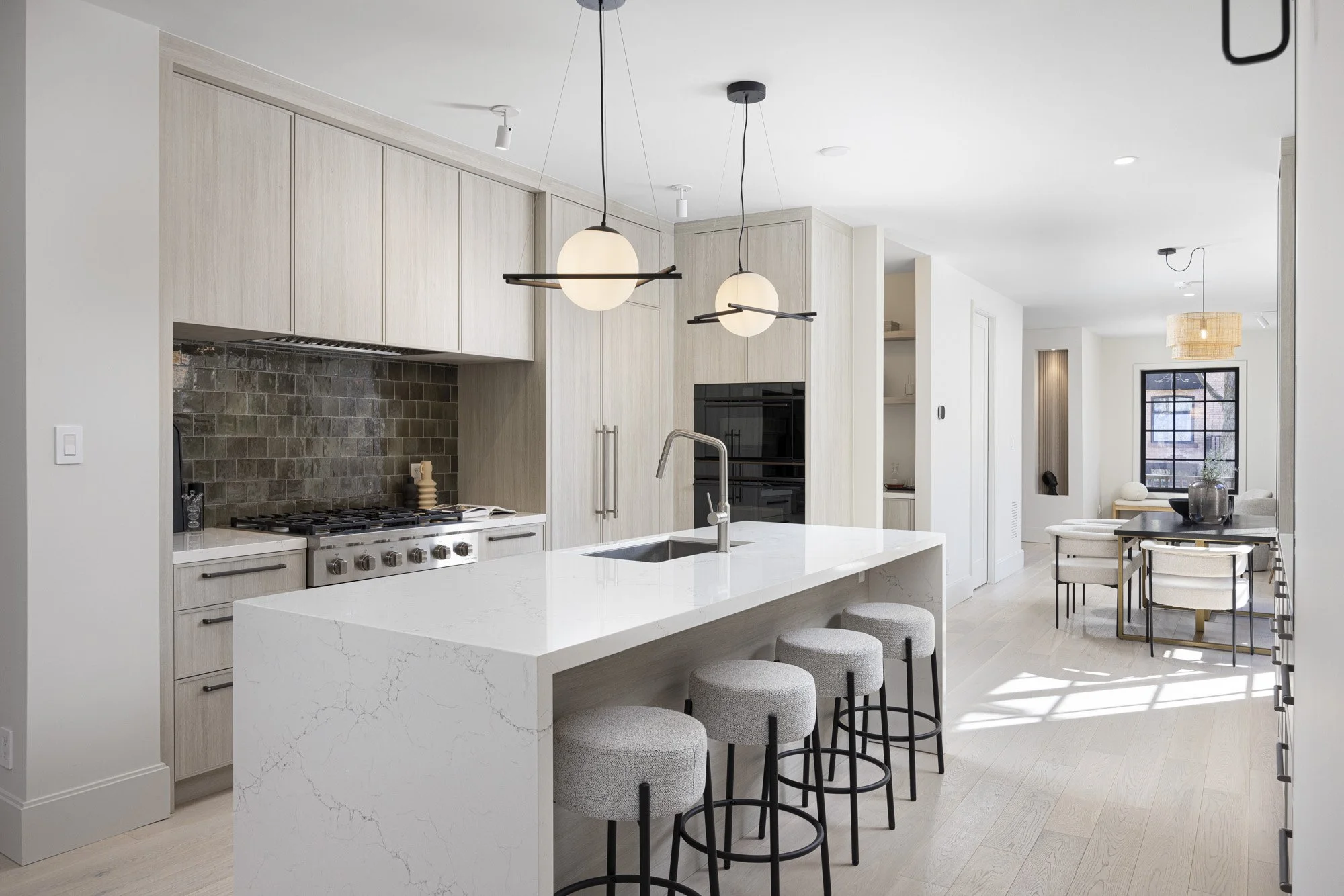 Modern open-concept kitchen and dining area with white marble island, light wood cabinetry, black tile backsplash, hanging pendant lights, and a round dining table with chairs.
