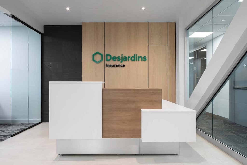 Reception desk in an office lobby with wood and white design, logo on the wall for Desjardins Insurance, glass walls, and modern lighting.