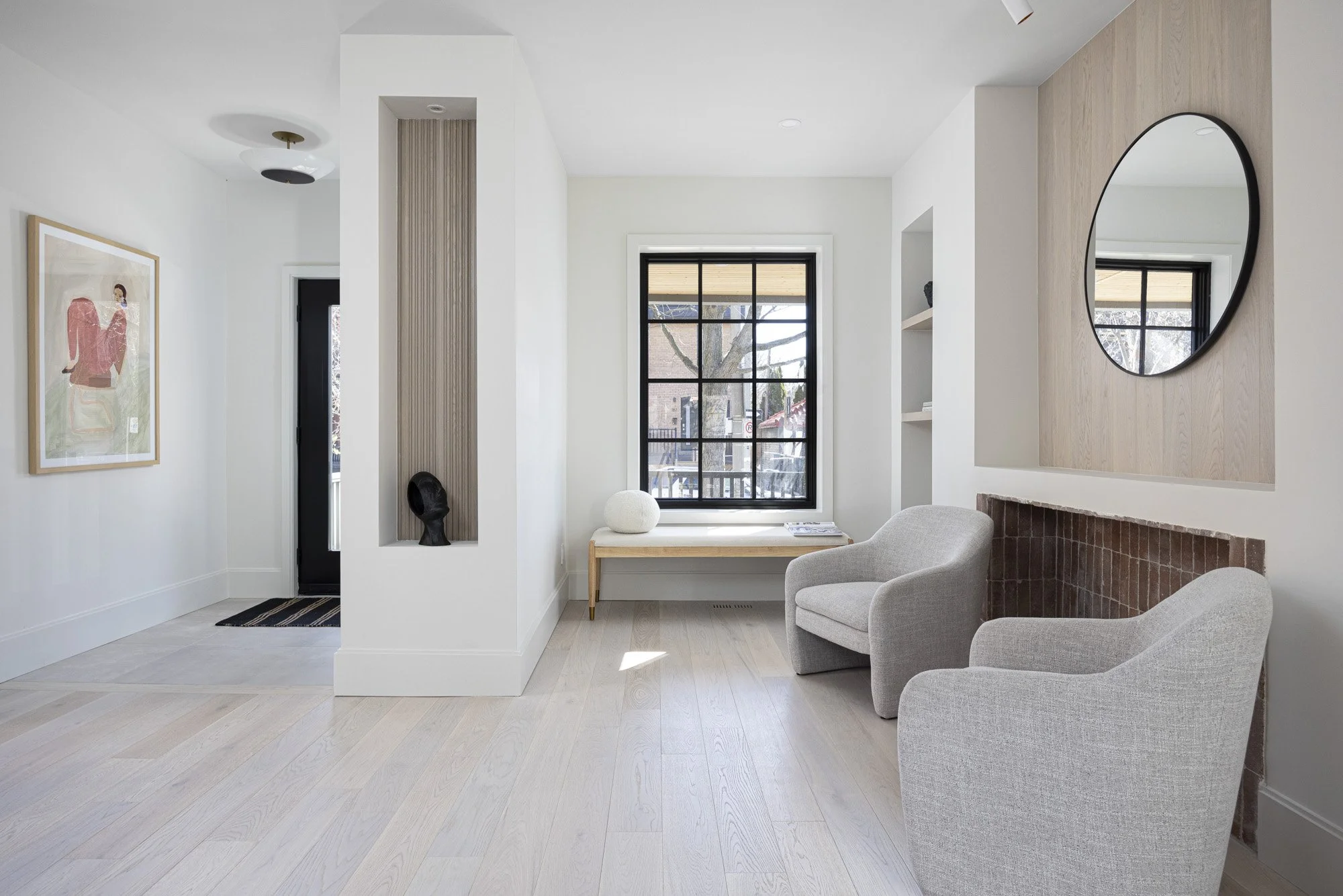 Modern living room with two light gray armchairs, a window with black frames, a wooden console table, and a round mirror on the wall.