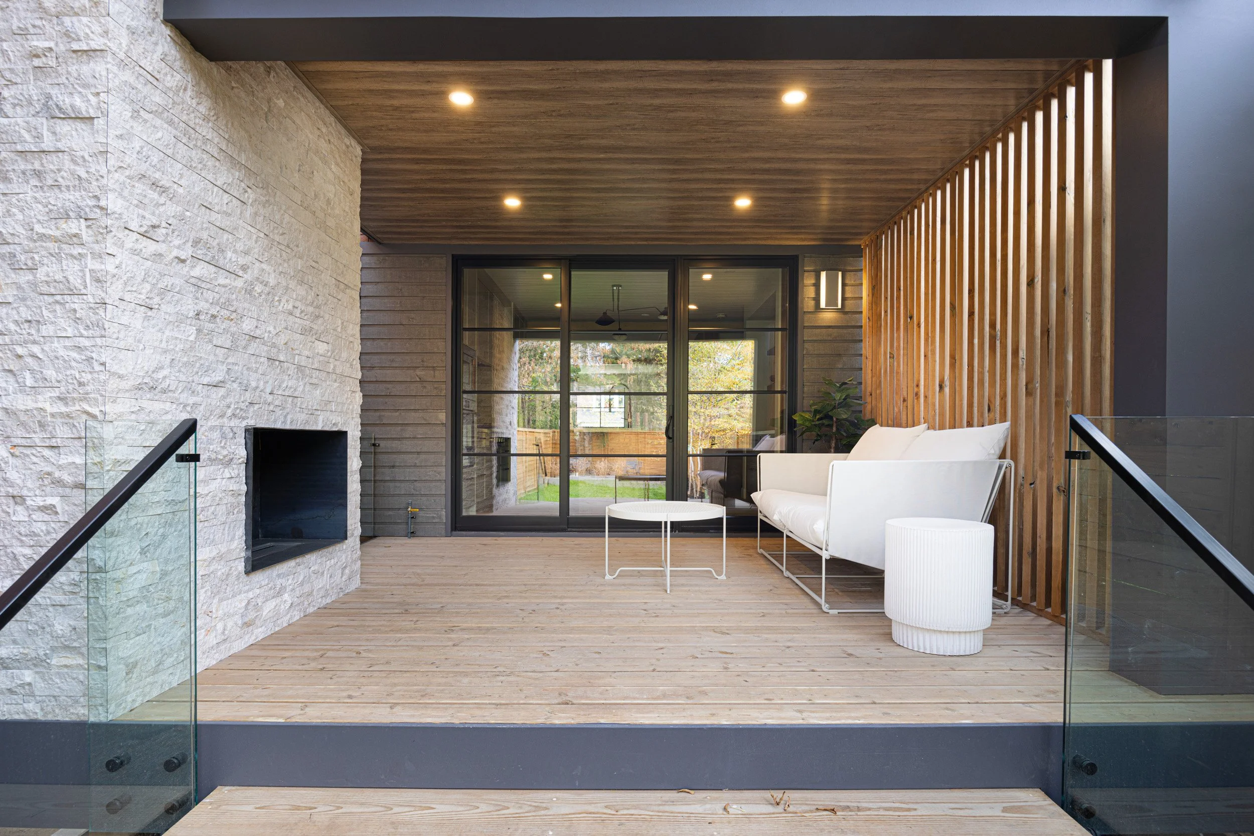 Modern outdoor patio with white furniture, glass railing, wood flooring, stone wall, and a wooden slat screen, with glass sliding doors leading into the house.
