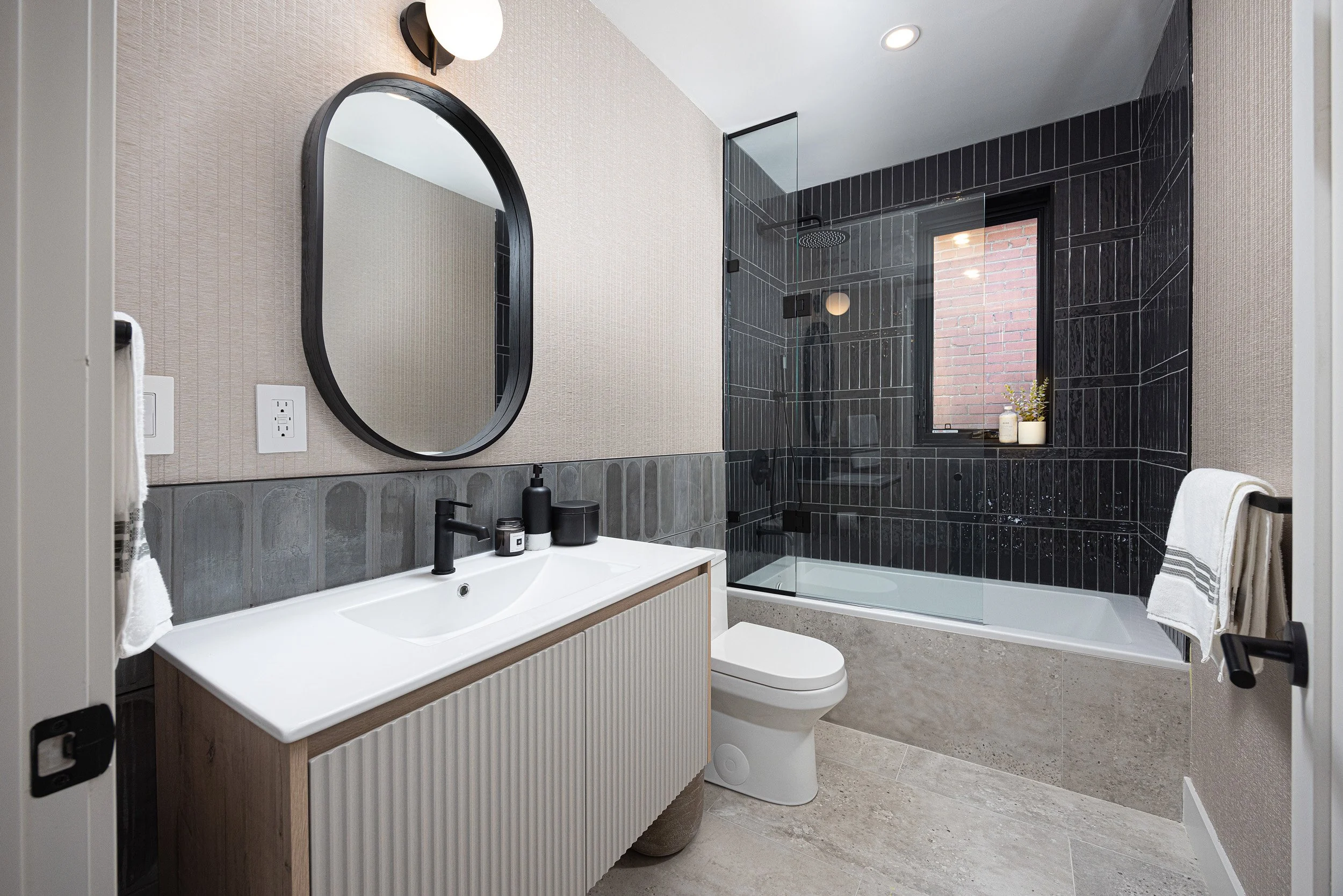 Modern bathroom with a white vanity, black faucet, oval mirror, black textured tiles in the shower area, a window with a brick wall outside, and towels on black towel bars.