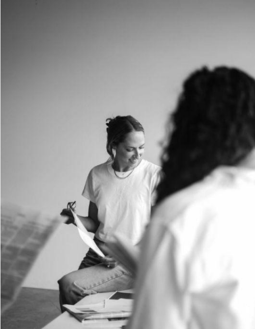 Two women sitting in an office or classroom, one holding a piece of paper and smiling, the other with curly hair, partially visible.