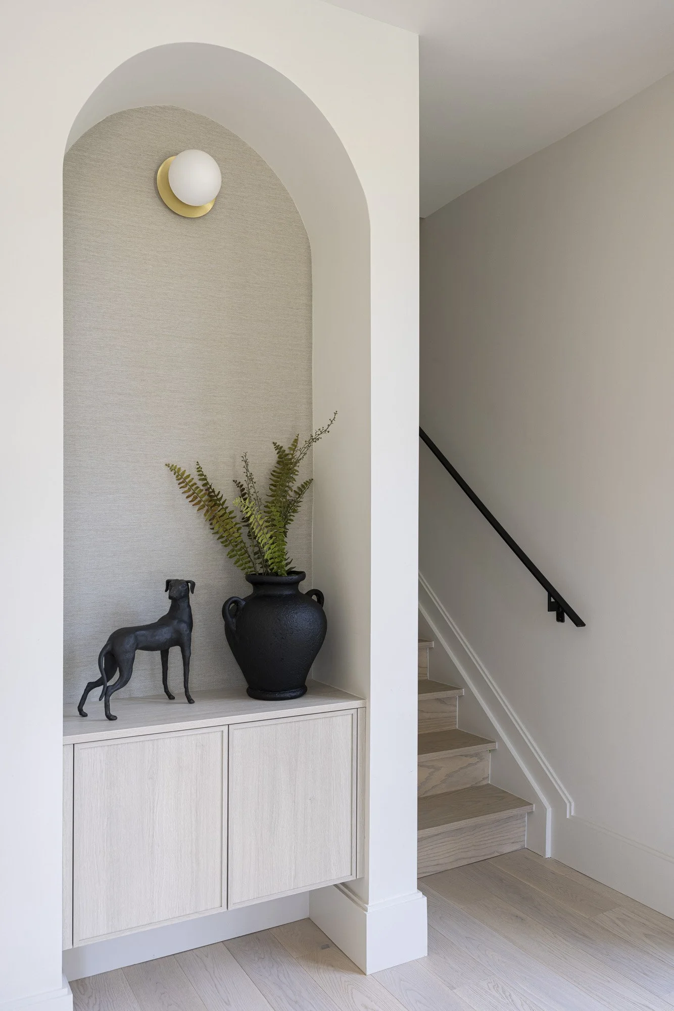 Decorative alcove with a black vase containing green ferns, a black dog figurine, and a circular wall light, next to a staircase with wooden steps and a black handrail.