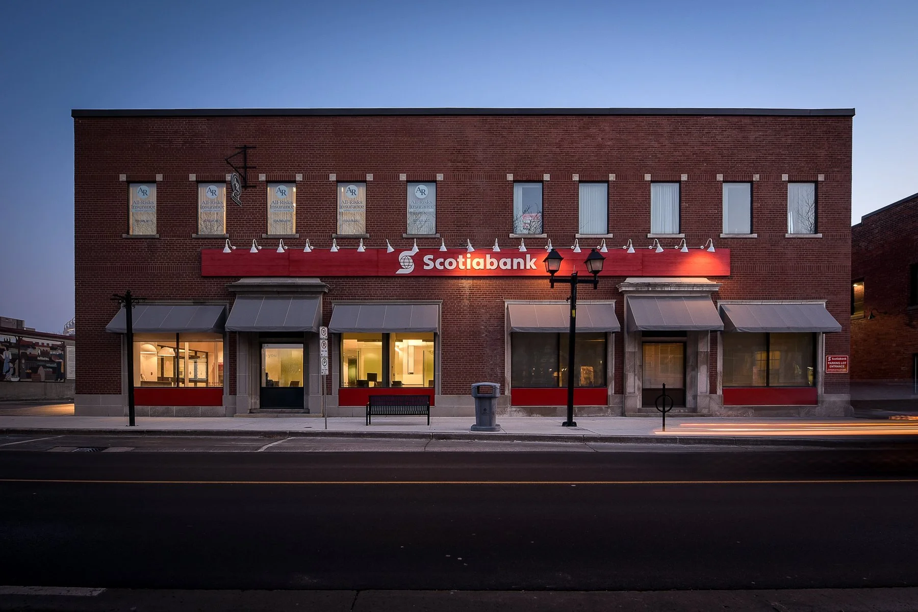 A red brick building housing a Scotiabank branch at dusk, with lit windows, awnings over the entrance, a bench, street lamps, and a trash can in front.