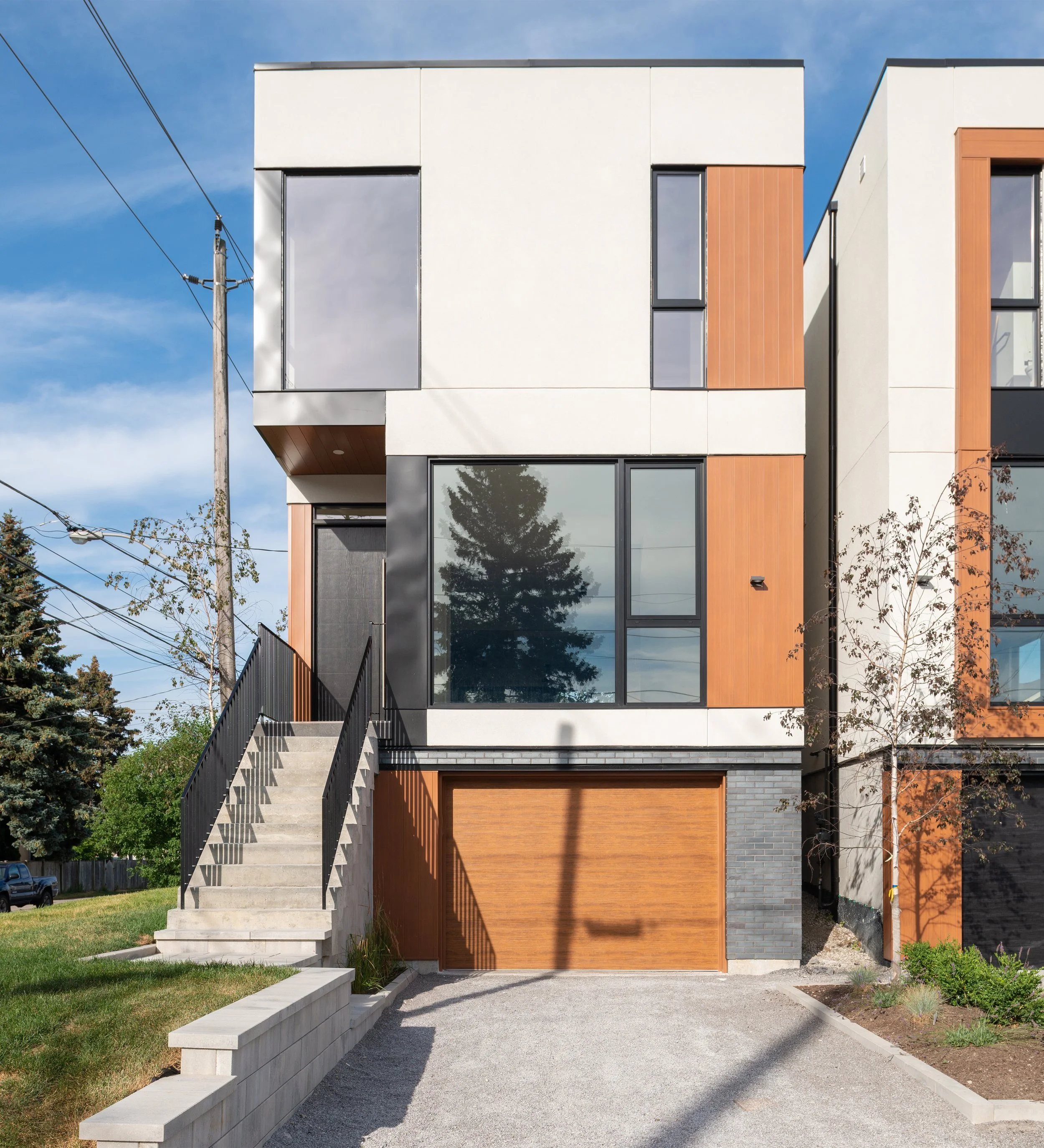 Modern multi-story house with a white and wood exterior, large front window, and a set of concrete stairs leading to the front door.