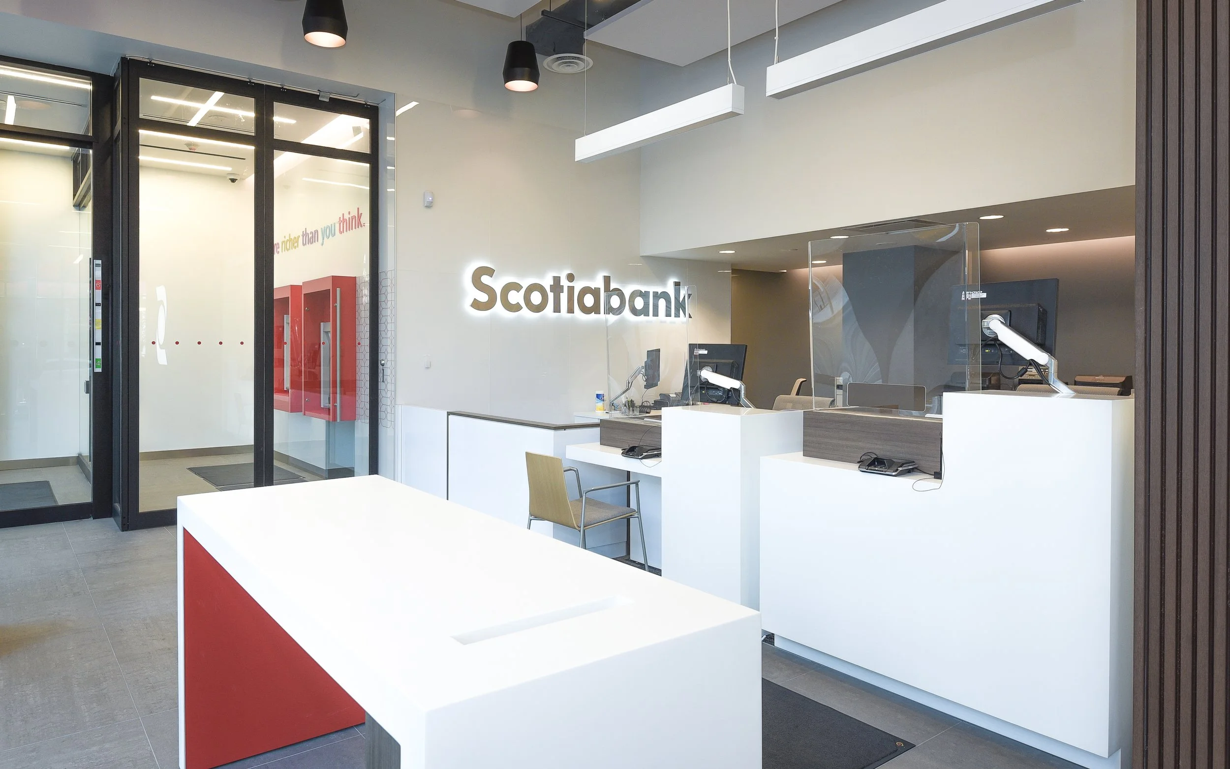 Scotiabank interior with a white counter, computers, a chair, and a glass partition. The bank's logo is illuminated on the wall, and there are black pendant lights hanging from the ceiling.