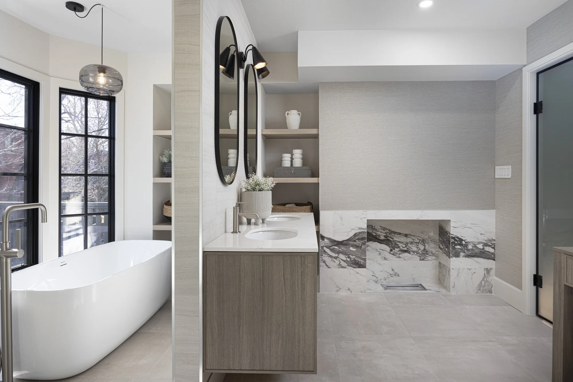 Modern bathroom with a freestanding bathtub near large windows, double sinks with mirrors, open shelves with decor, and a marble-tiled walk-in shower.