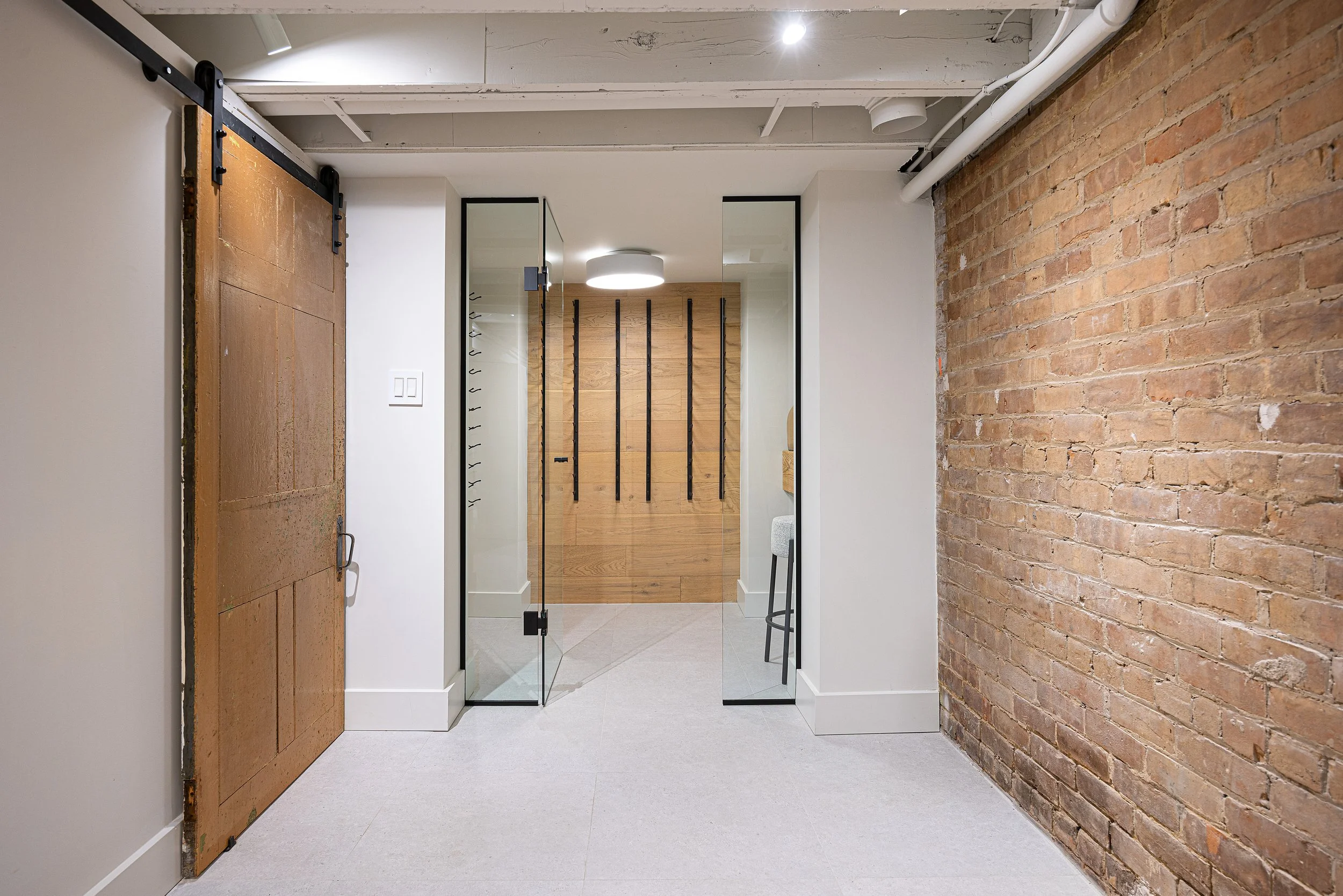 Interior of a modern basement with a brick wall on the right, glass doors in the center with black metal frames, a wooden wall in the background with black hooks, and a sliding barn door on the left.