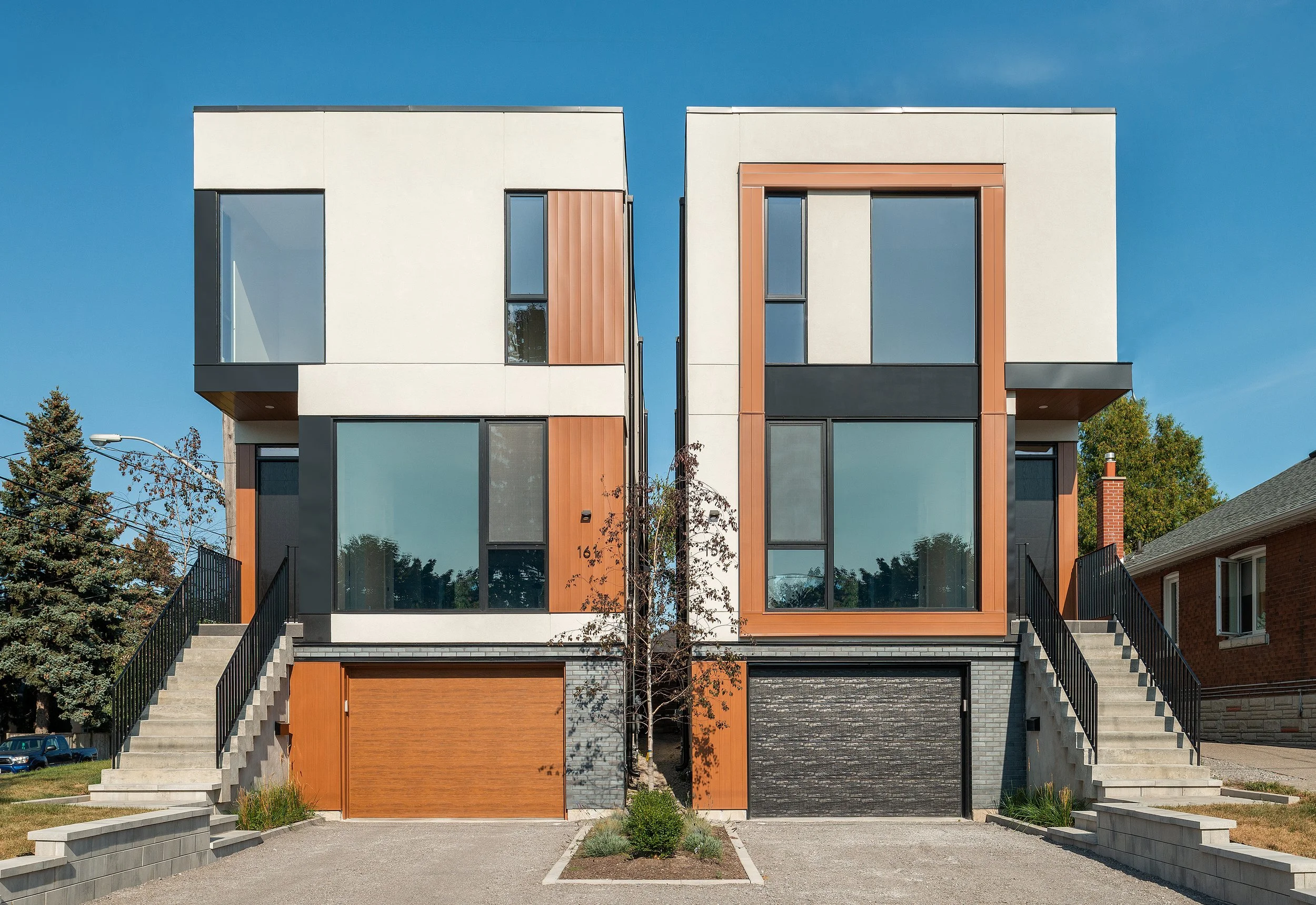 Modern duplex with symmetrical design, large glass windows, wooden accents, concrete stairs, and garages in front, under a clear blue sky.