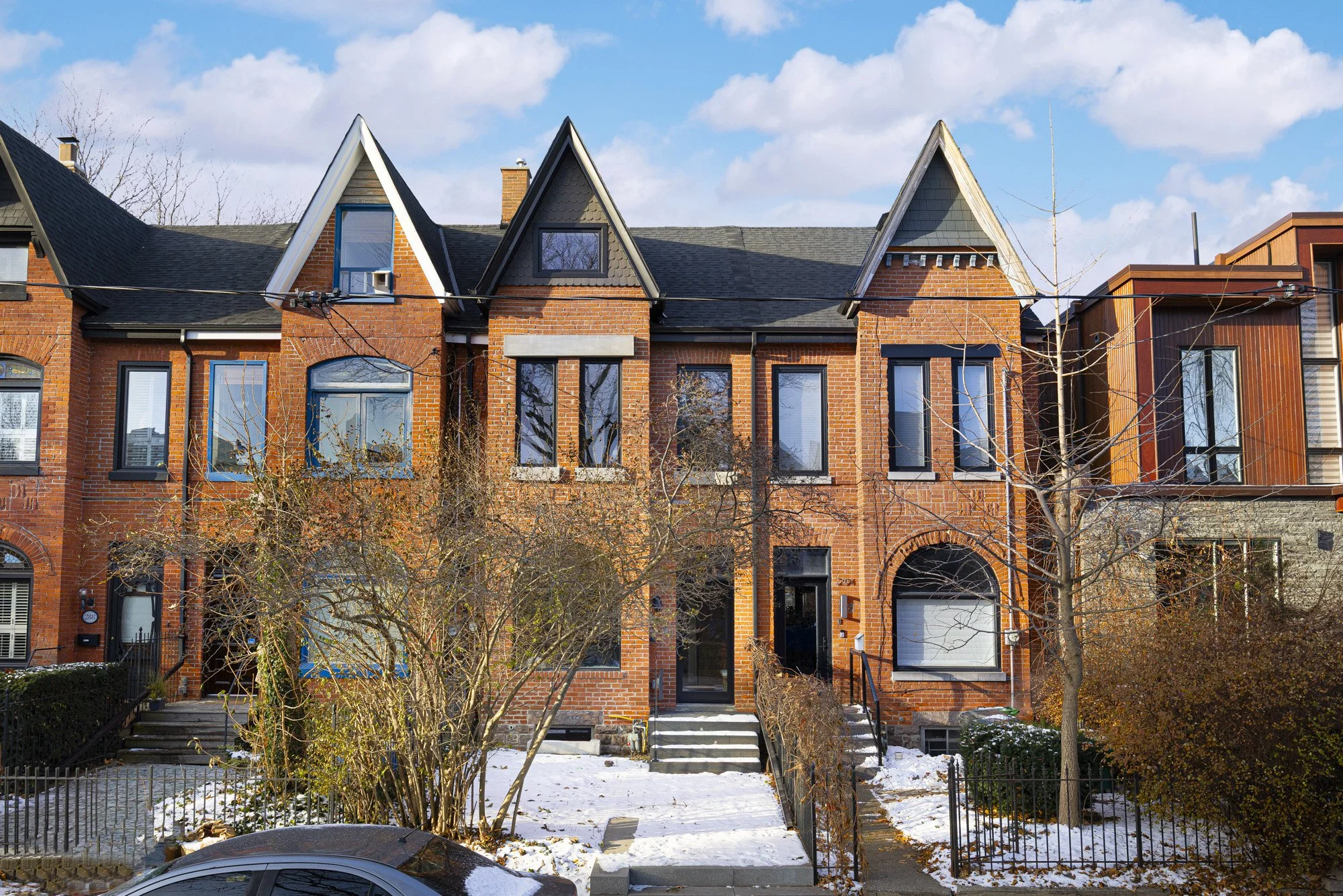 Row of brick residential houses with large windows, steps leading to entrances, and a snow-covered front yard with trees and bushes.