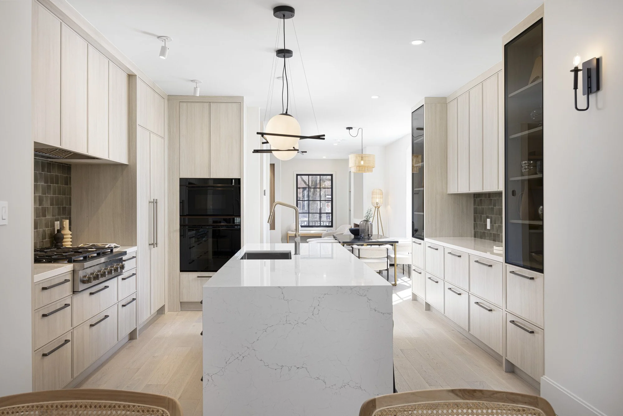 Modern kitchen with light wood cabinets, white marble island, black appliances, and minimalist lighting fixtures, opening to a dining and living area with large window.