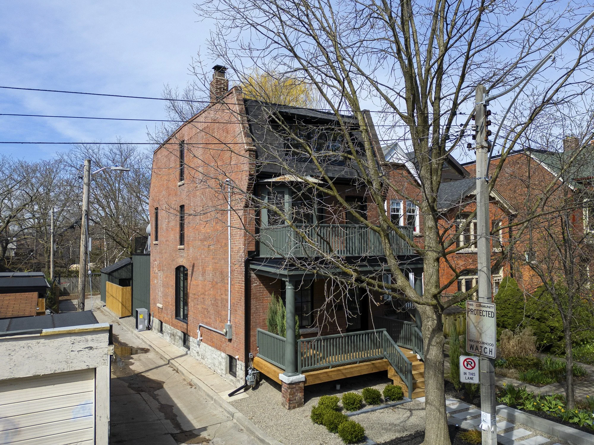 A three-story brick house with a balcony and front porch, leafless trees, utility poles, and a street sign reading 'Protected Neighborhood Watch' in front.