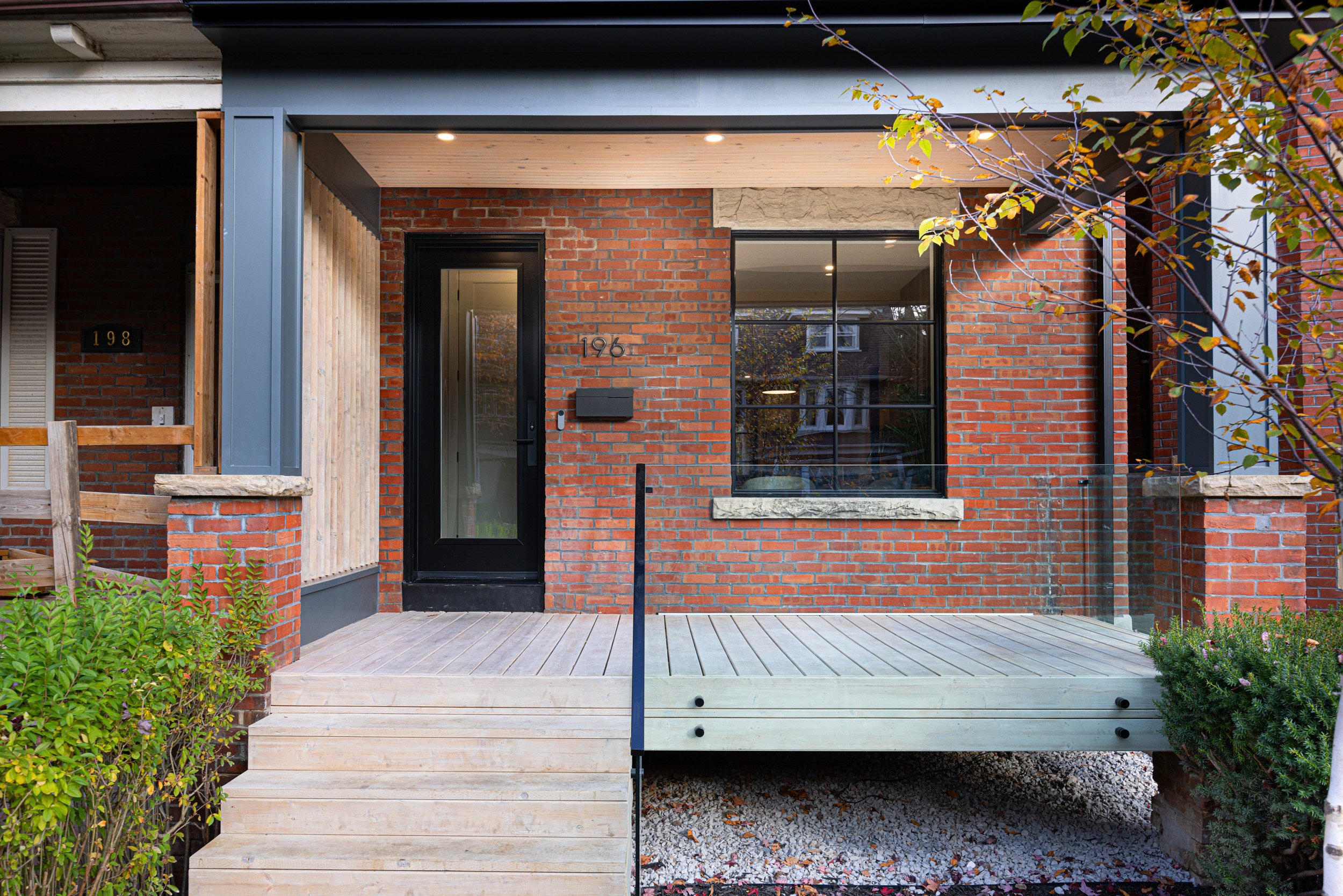Newly renovated brick house with a wooden front porch, black door, and large window, surrounded by greenery.