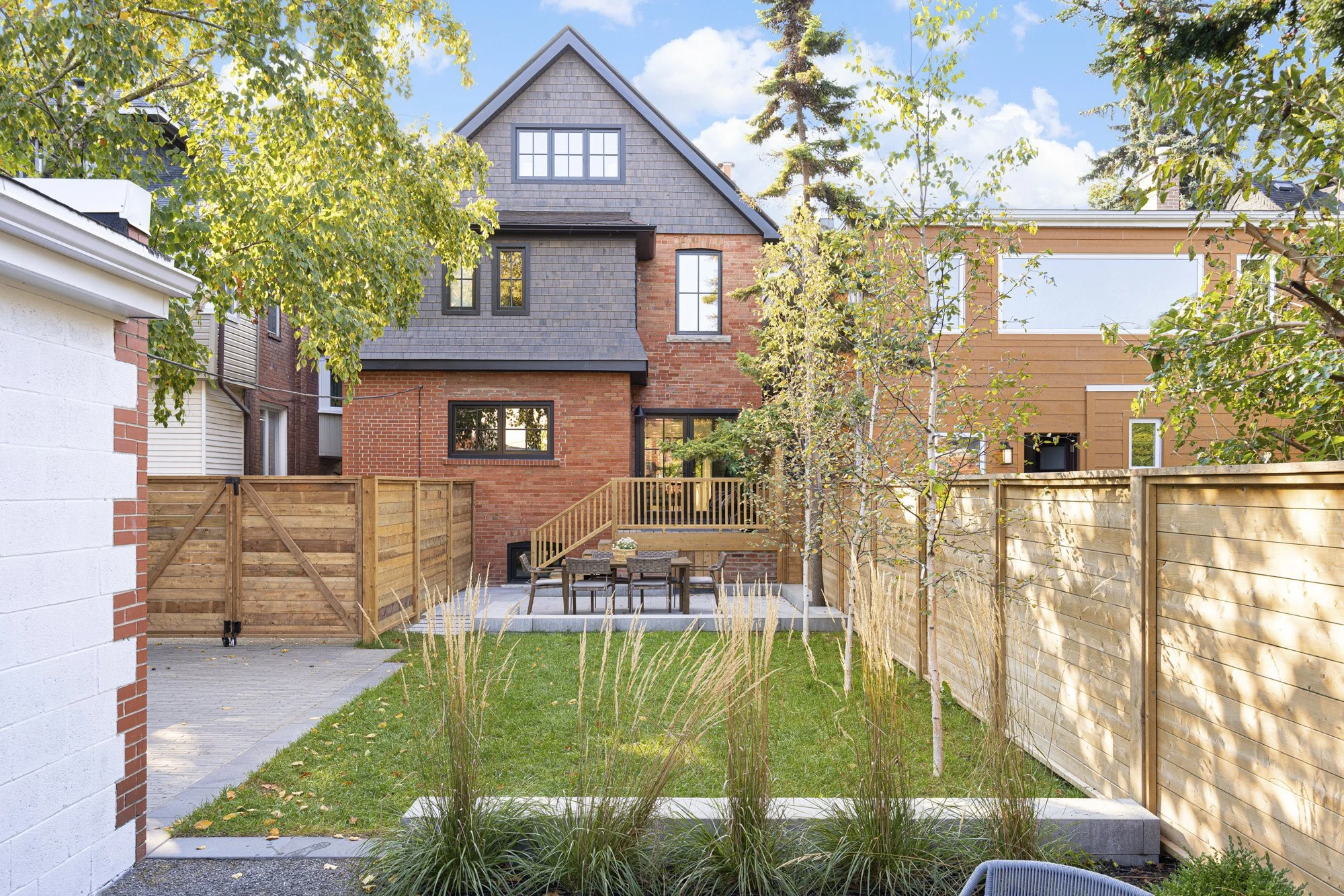 A backyard with a grassy lawn, a wooden fence, a small patio with a table and chairs, a deck with a railing, trees, and neighboring houses with brick and wood exteriors under a partly cloudy sky.