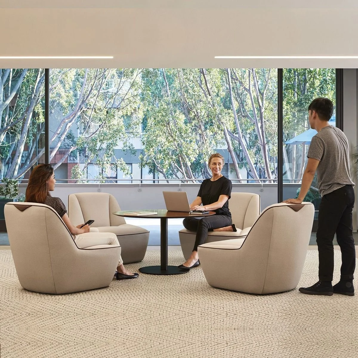 Three people in a modern office lounge area with large windows showing trees outside. Two women are seated, one with a smartphone and the other with a laptop, while a man stands and leans on a chair, engaged in conversation.