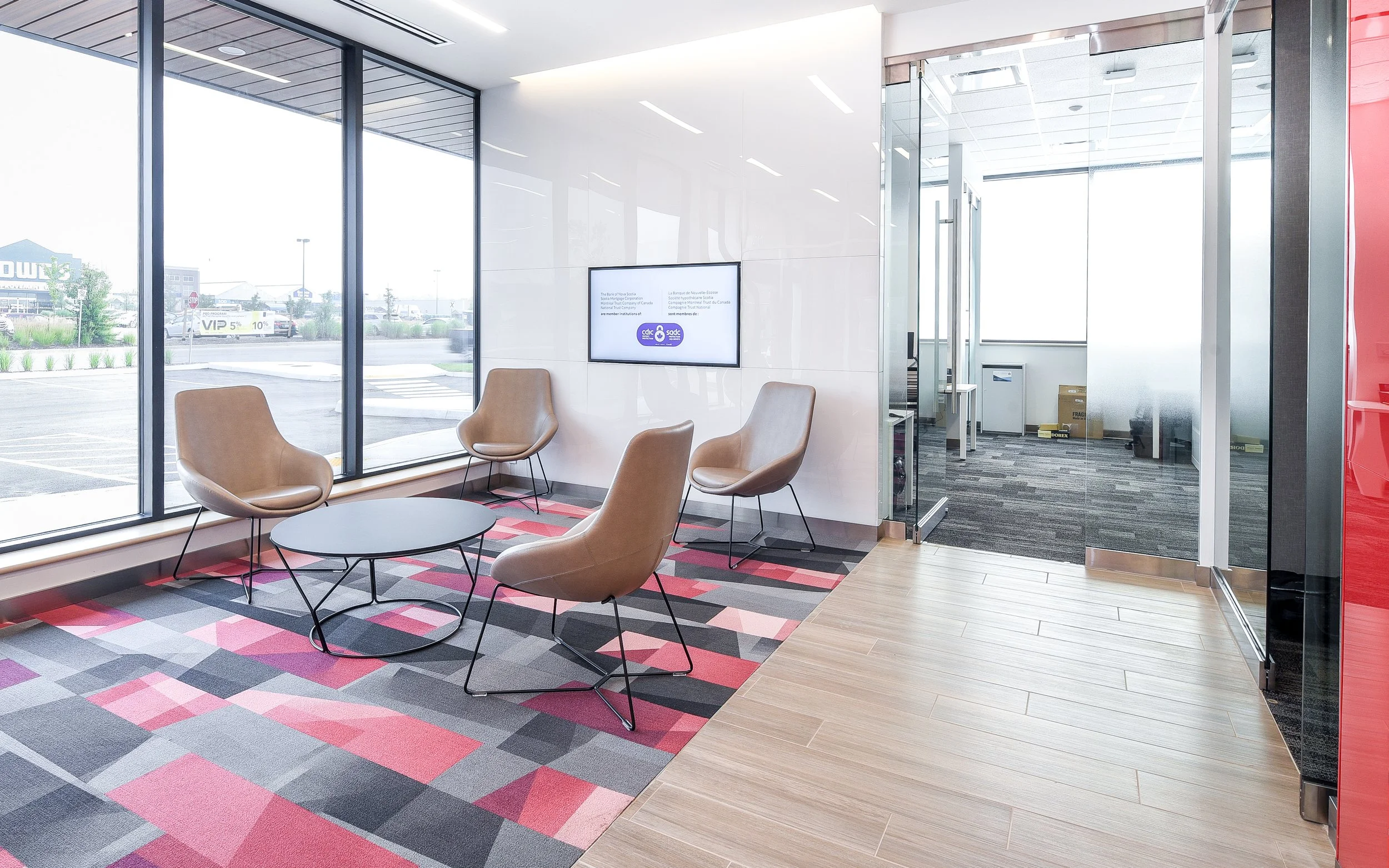 Modern office waiting area with four tan chairs, a small round black coffee table, floor-to-ceiling windows, a wall-mounted TV, and a separate conference room with frosted glass doors visible in the background.