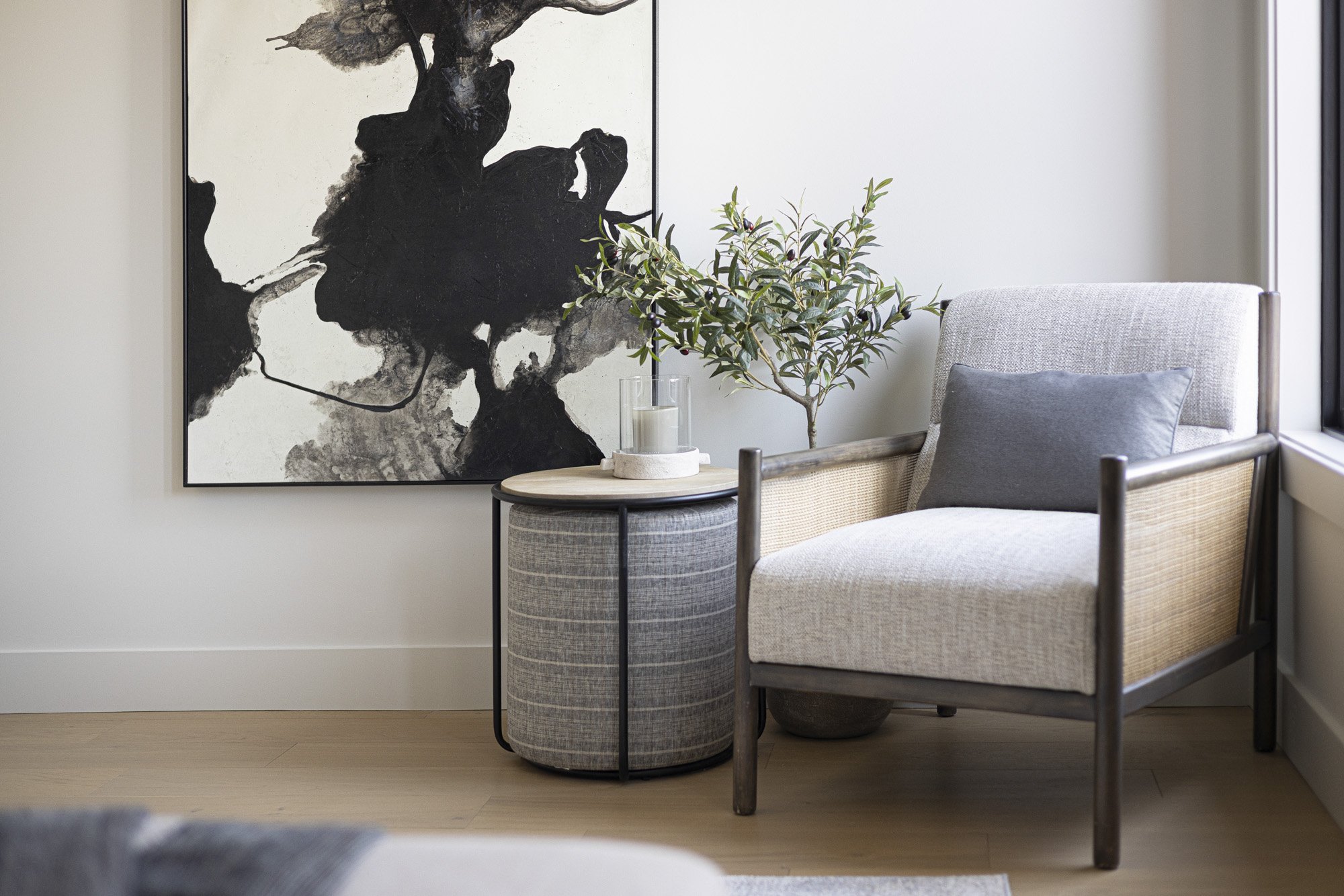 A cozy corner with a beige armchair that has dark wooden arms and legs, a gray pillow on the seat, and a large abstract black-and-white painting on the wall behind it. A small round side table with a light wooden top, black metal frame, and gray fabric-covered cylindrical base is next to the chair. On the table is a glass container holding a white candle and a green leafy plant in a pot.