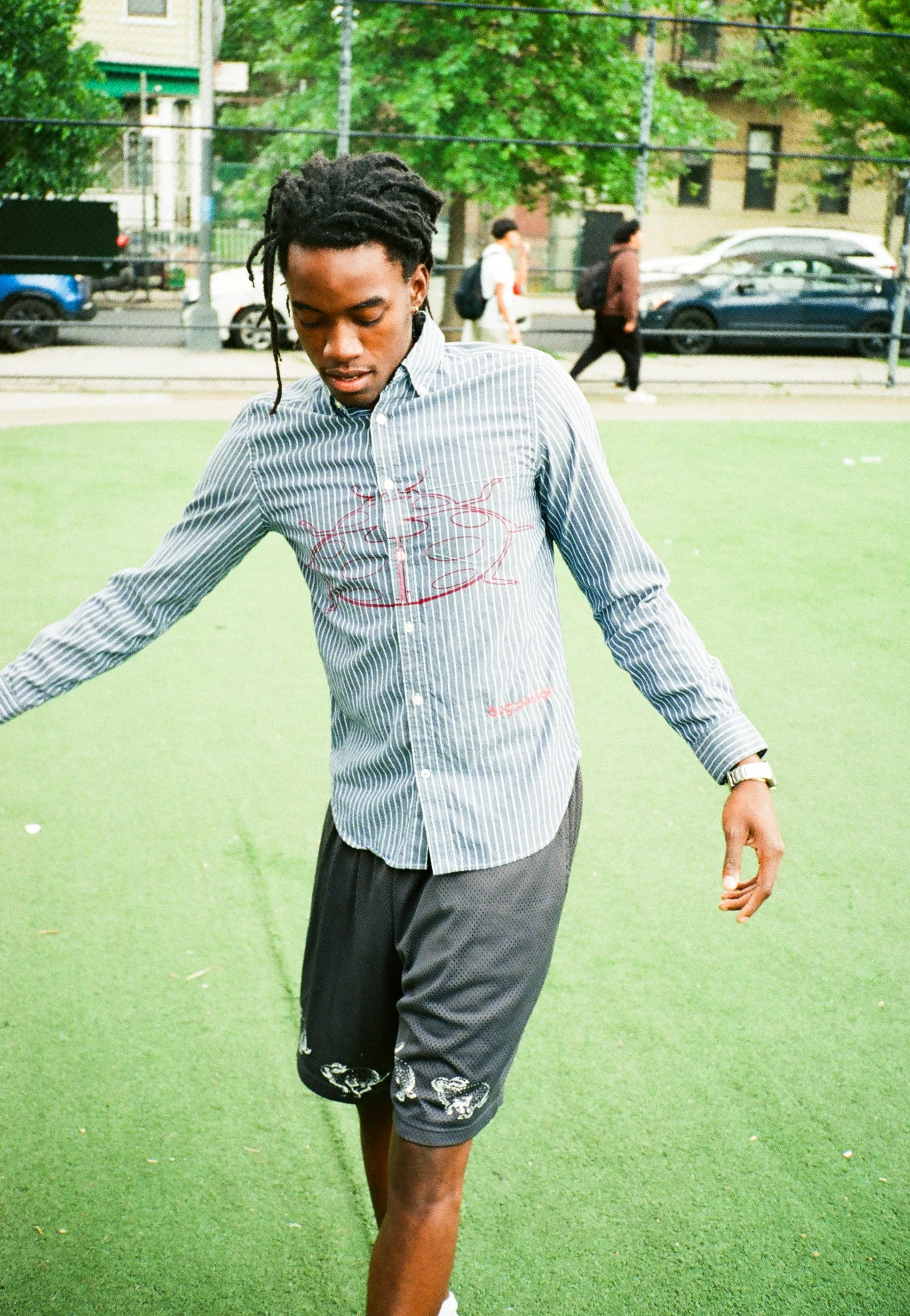 A young man with dreadlocks and a striped shirt walking on a green artificial turf field in an urban park with trees and a metal fence behind him. In the background, other people walk along the sidewalk beside parked cars and buildings.
