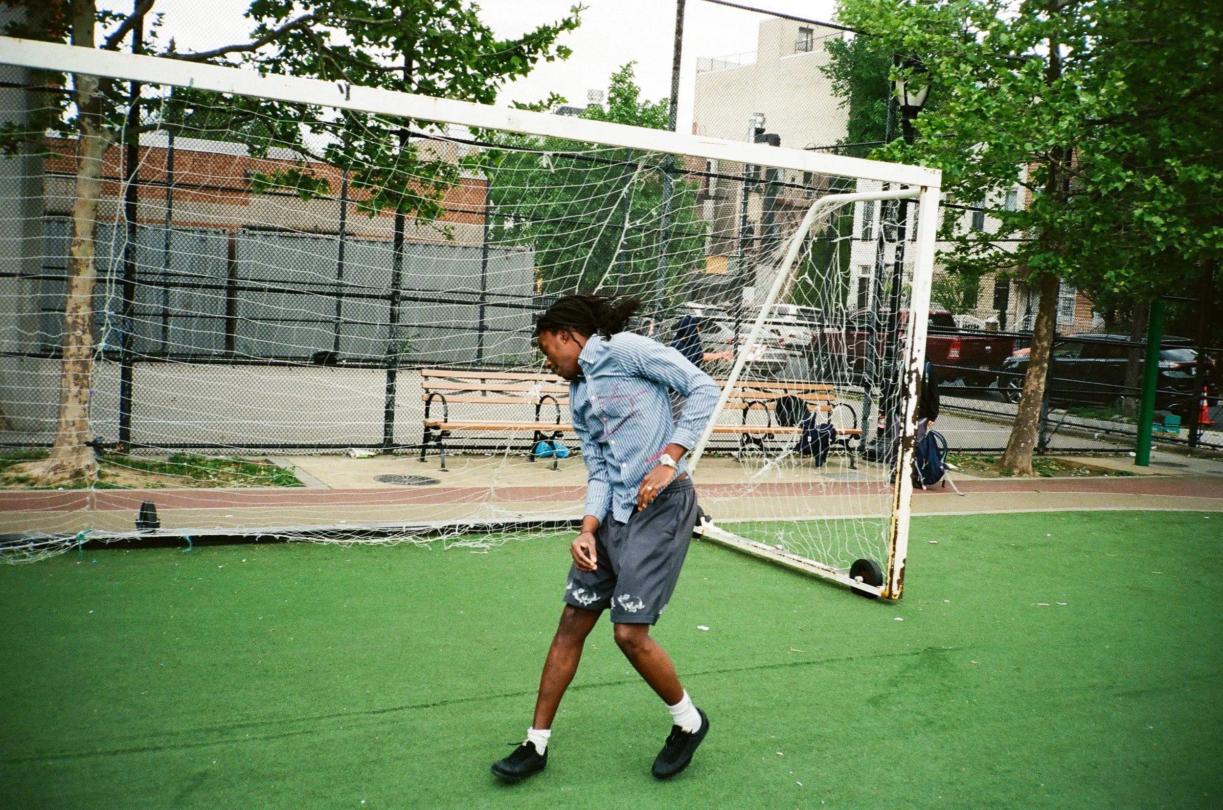 A person with dreadlocks wearing a blue striped shirt and shorts walks past a broken soccer goal on an outdoor sports court with green artificial turf, trees, parked cars, and buildings in the background.