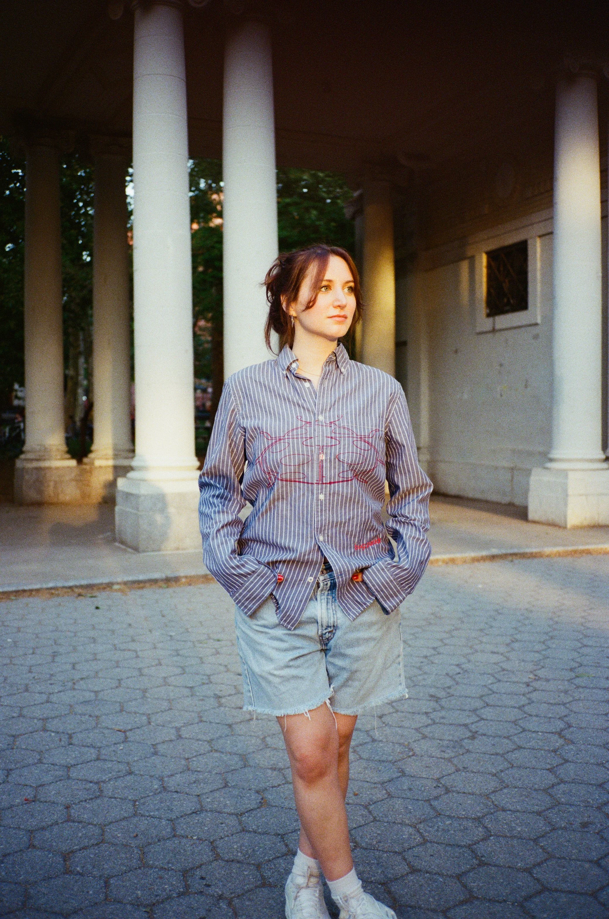 A young woman with shoulder-length brown hair standing outdoors on a cobblestone path in front of a building with tall white columns. She is wearing a striped button-up shirt with red embroidery, denim shorts, white socks, and sneakers. The lighting 