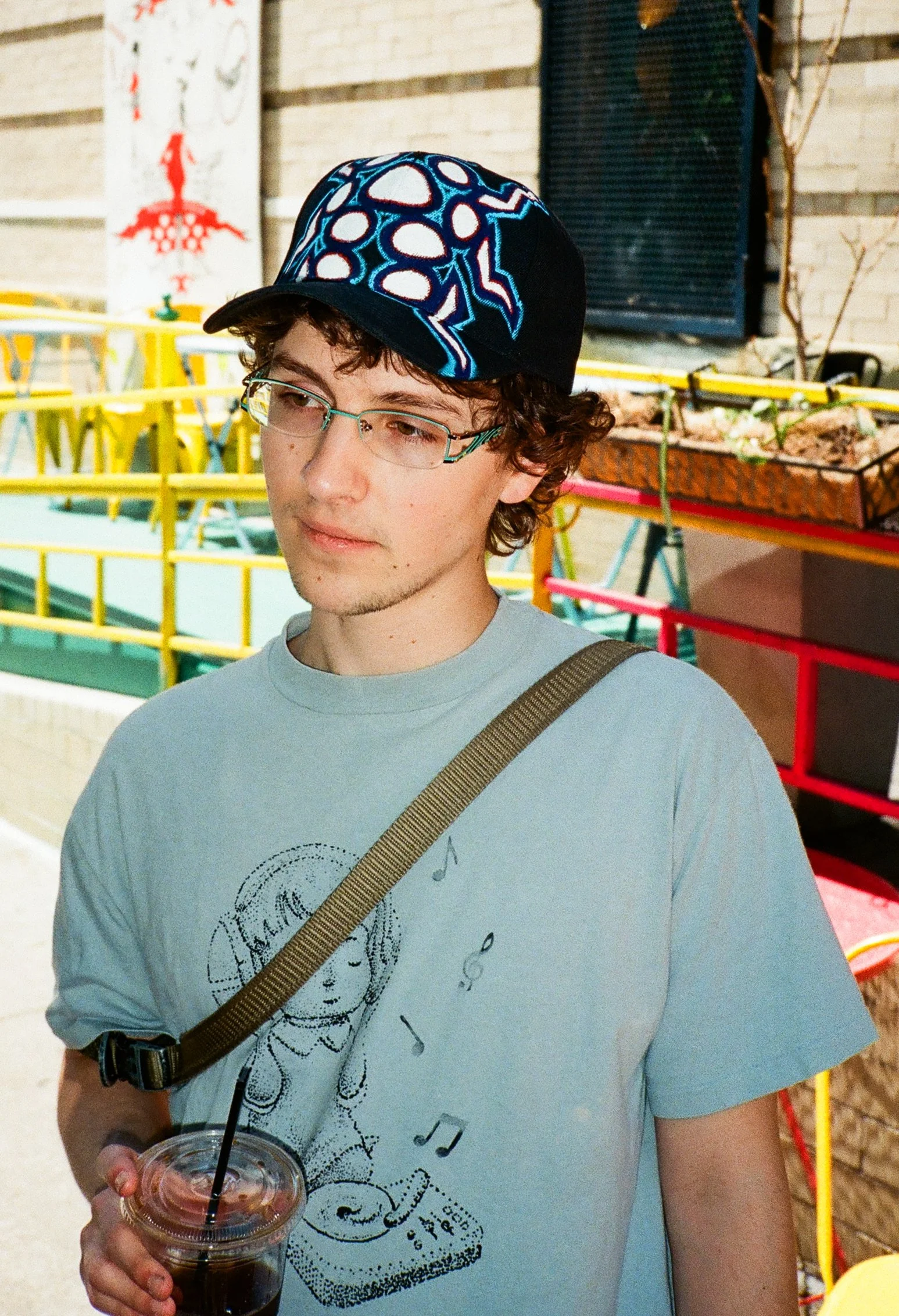 A young man with curly hair and glasses, wearing a black cap with blue and white patterns, holding a plastic cup of dark soda with a straw. He is outdoors near a brick wall and colorful metal railing.