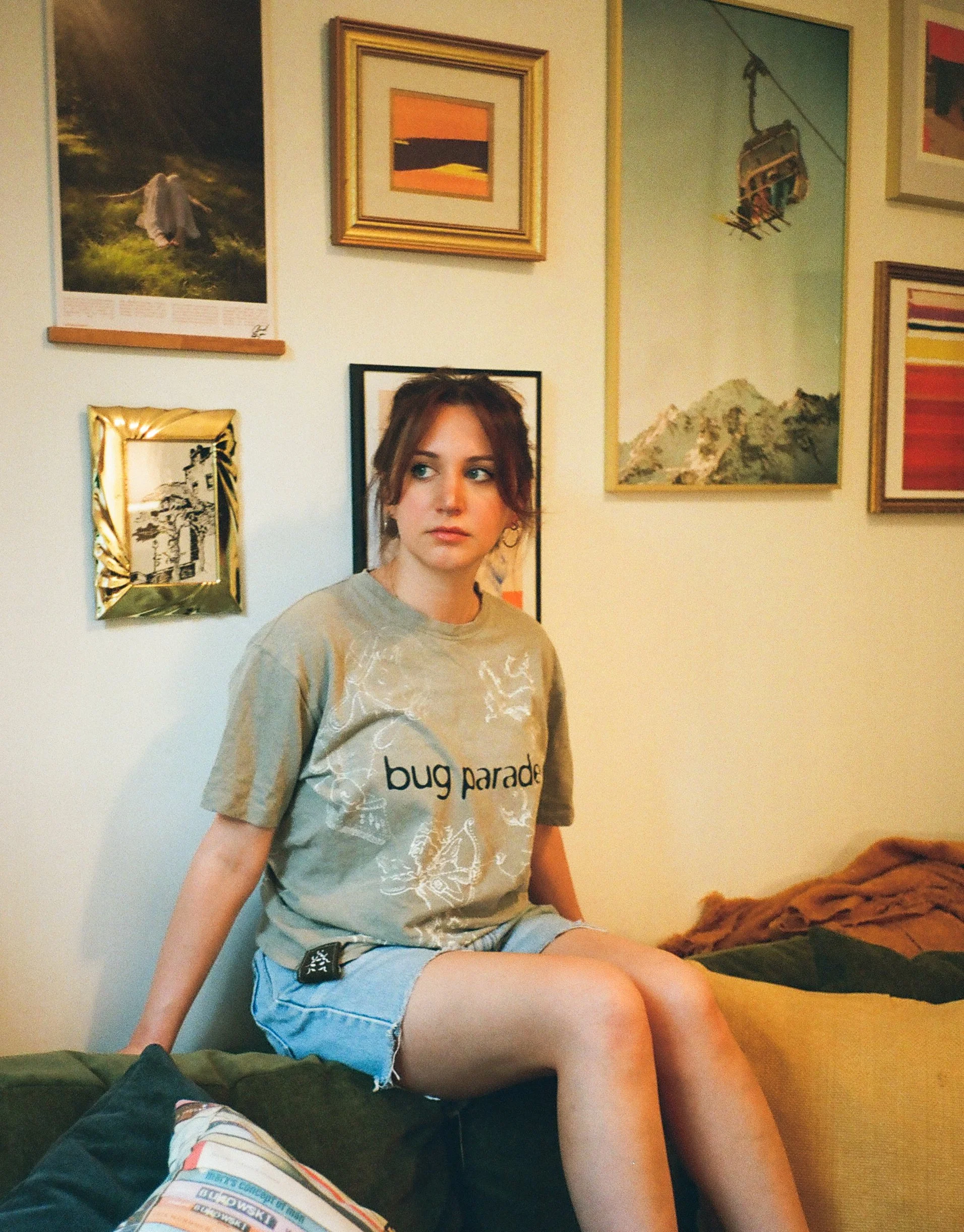 A young woman with reddish-brown hair, wearing a gray T-shirt with 'bug parade' written on it, sitting on a couch or bed, with a mountain landscape painting and other artwork on the wall behind her.