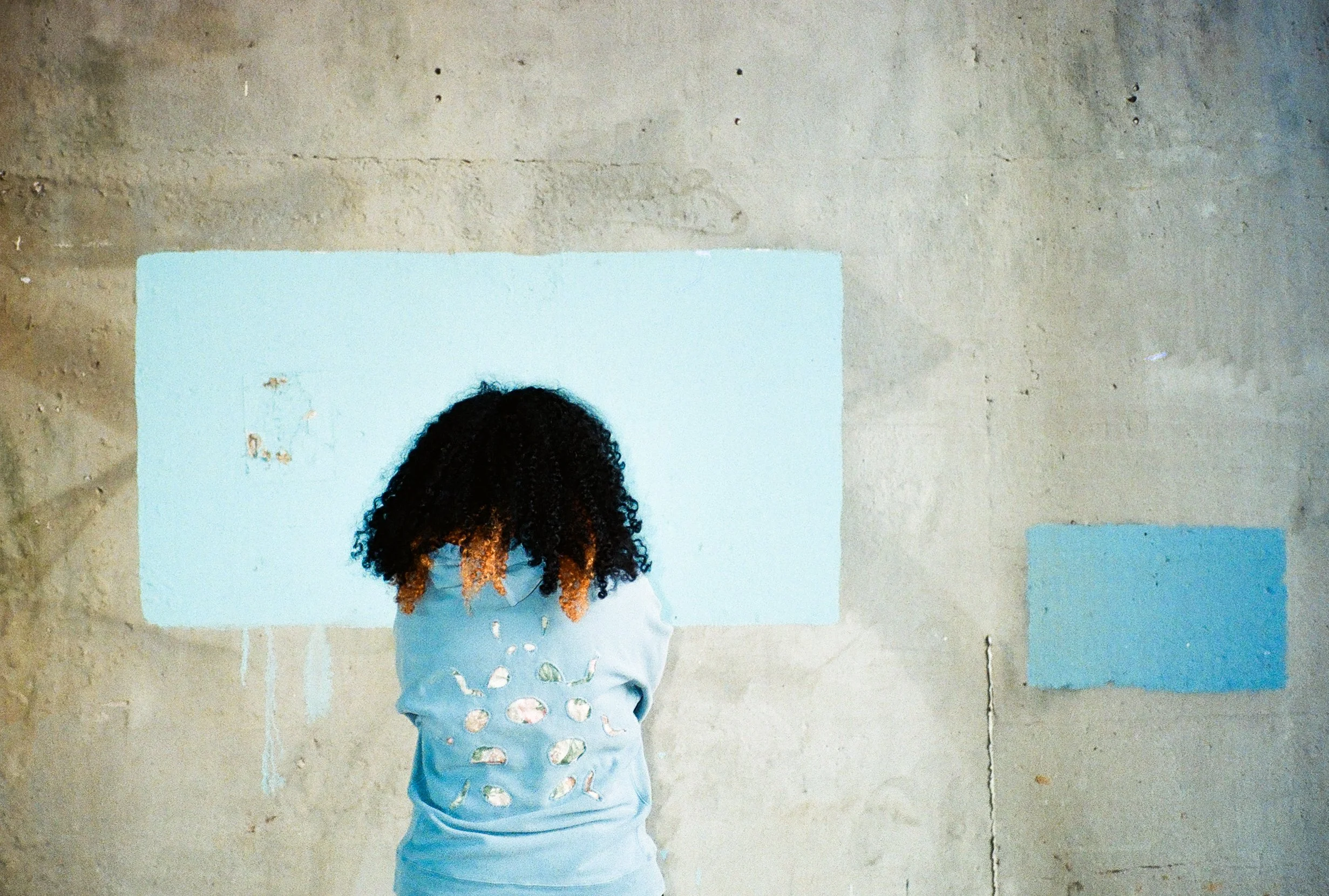 A girl with curly hair wearing a light blue shirt with cutouts on the back, standing in front of a plain, textured concrete wall with blue paper or posters on it.