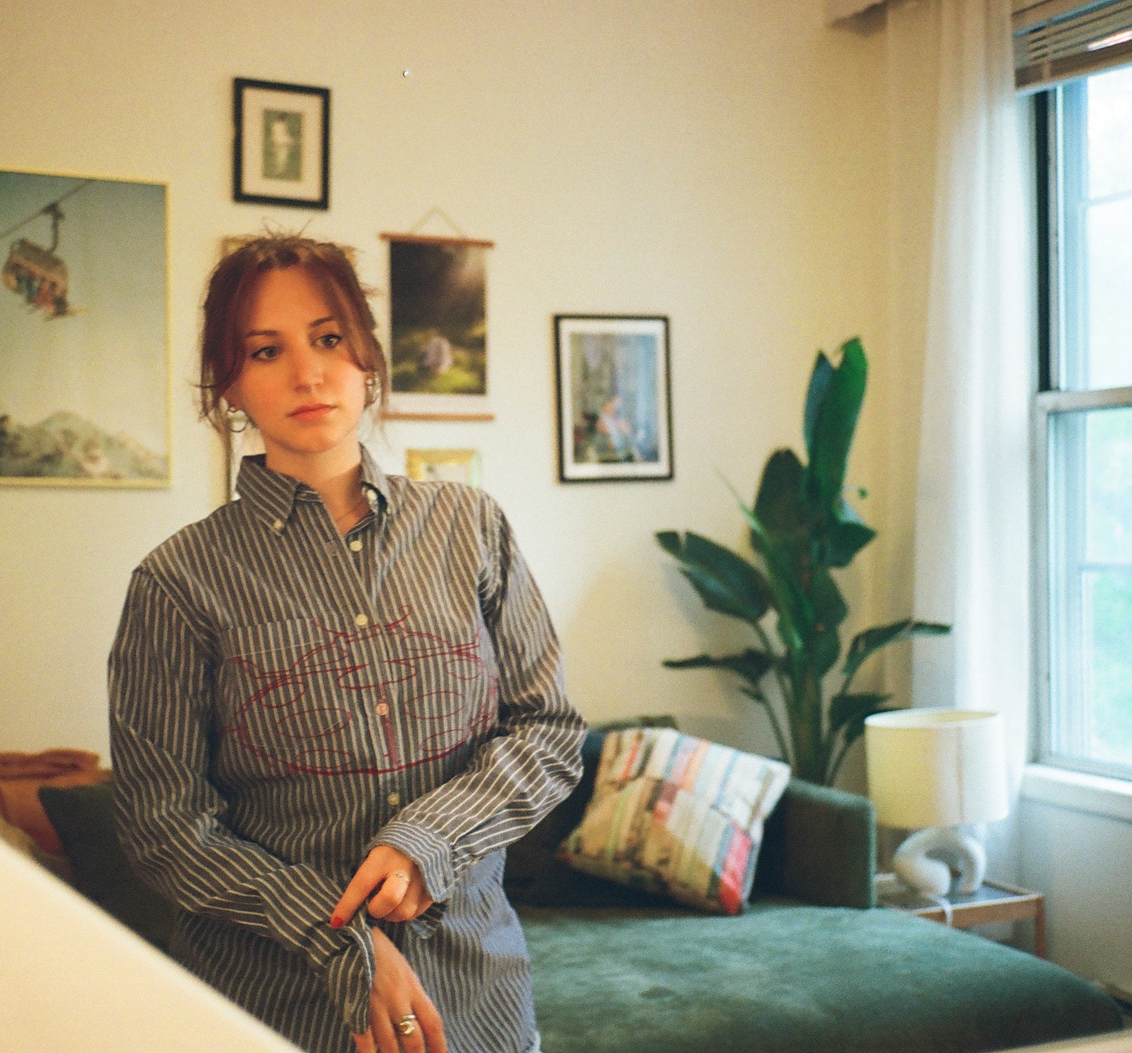 A young woman with reddish-brown hair, wearing a striped button-up shirt, standing indoors near a window with white curtains. The room has several framed pictures on the wall, a large green plant, and a bookshelf with colorful books. She appears to b