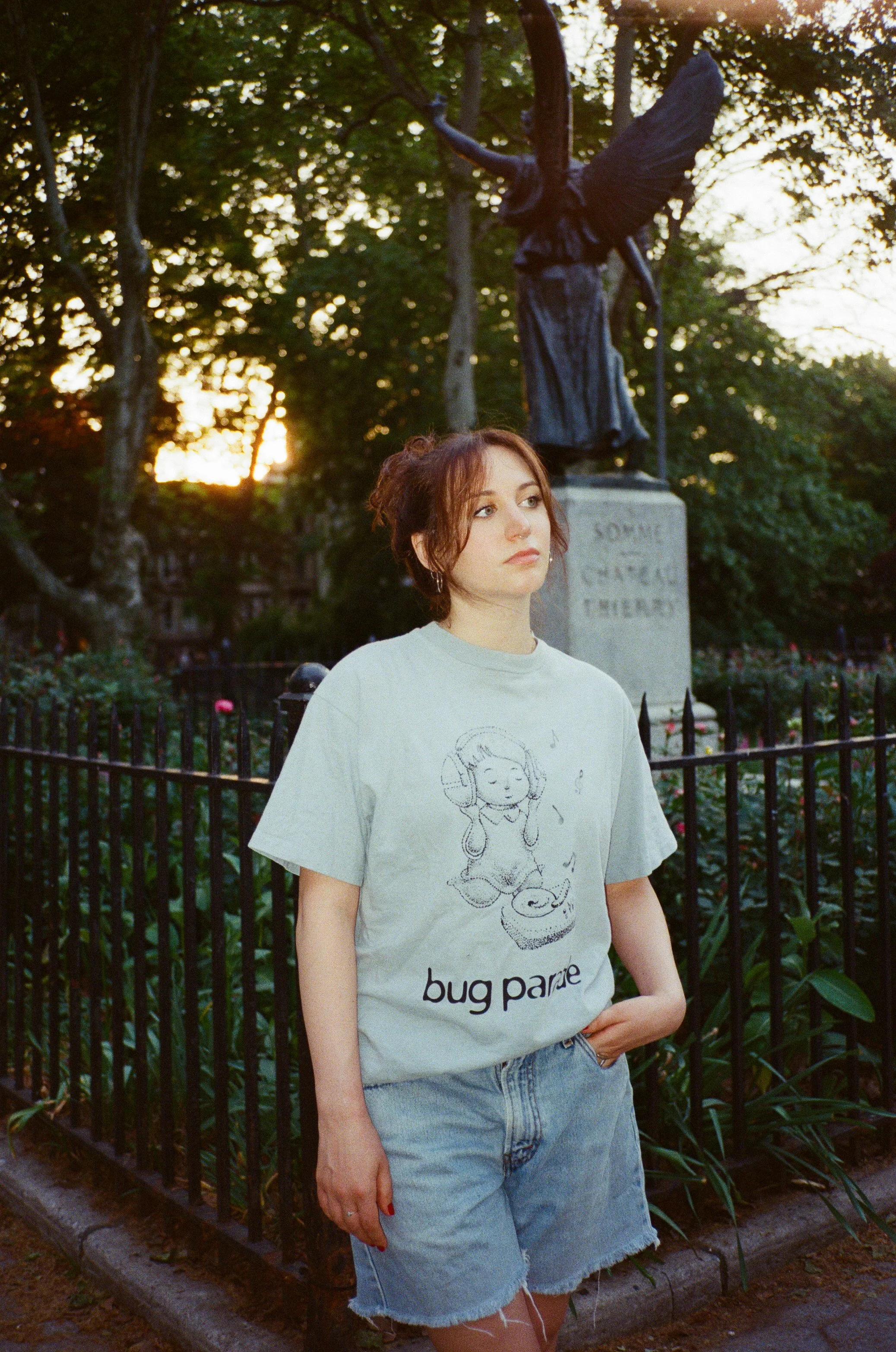 A young woman with red hair and a nose ring stands in front of a black iron fence and a statue of an angel, in a park during sunset, wearing a white graphic T-shirt with a cartoon girl and the words 'bug peace' and denim shorts.