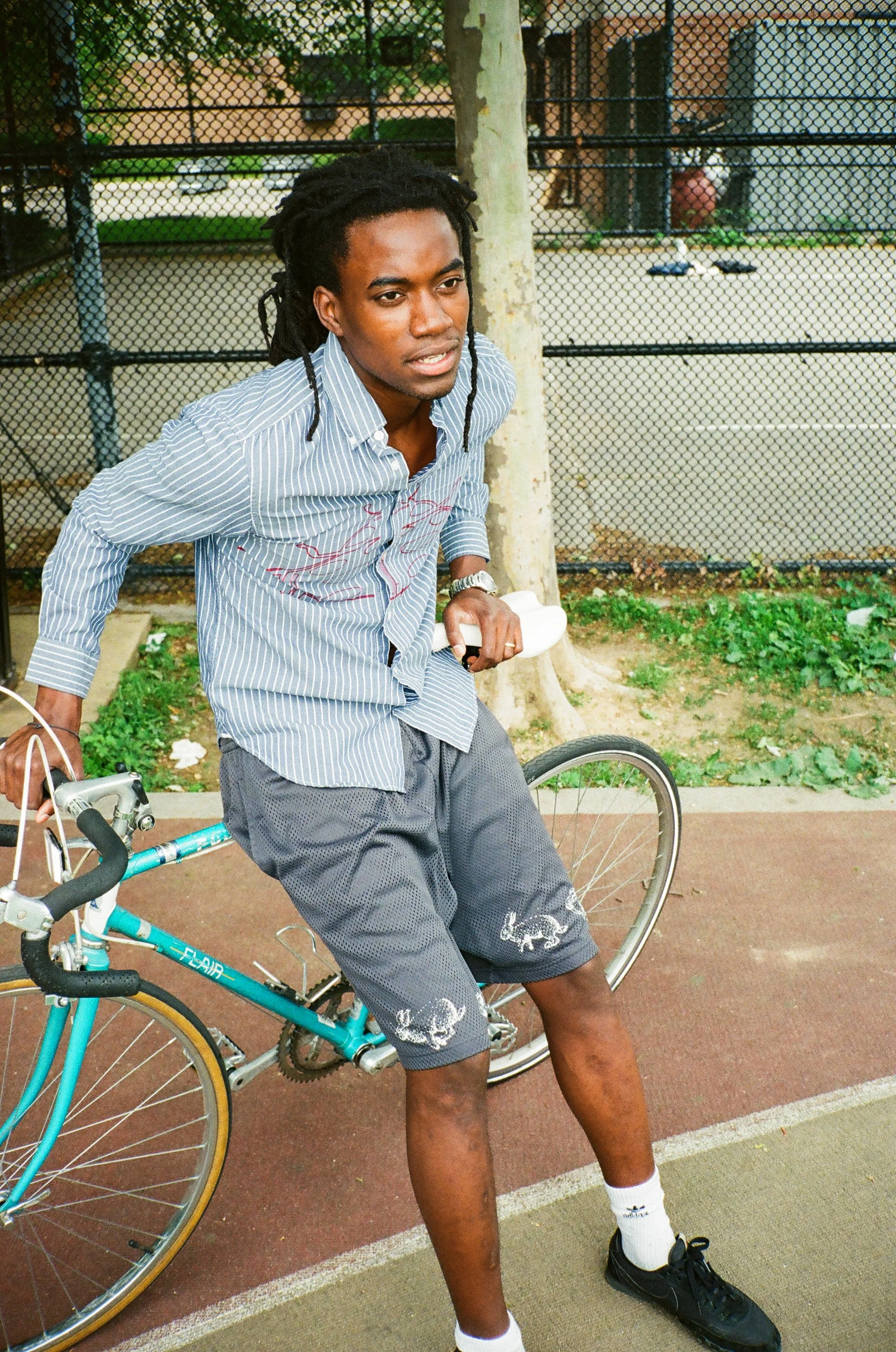 A young man with dreadlocks in a striped shirt and shorts standing next to a bicycle at an outdoor basketball court surrounded by a chain-link fence.