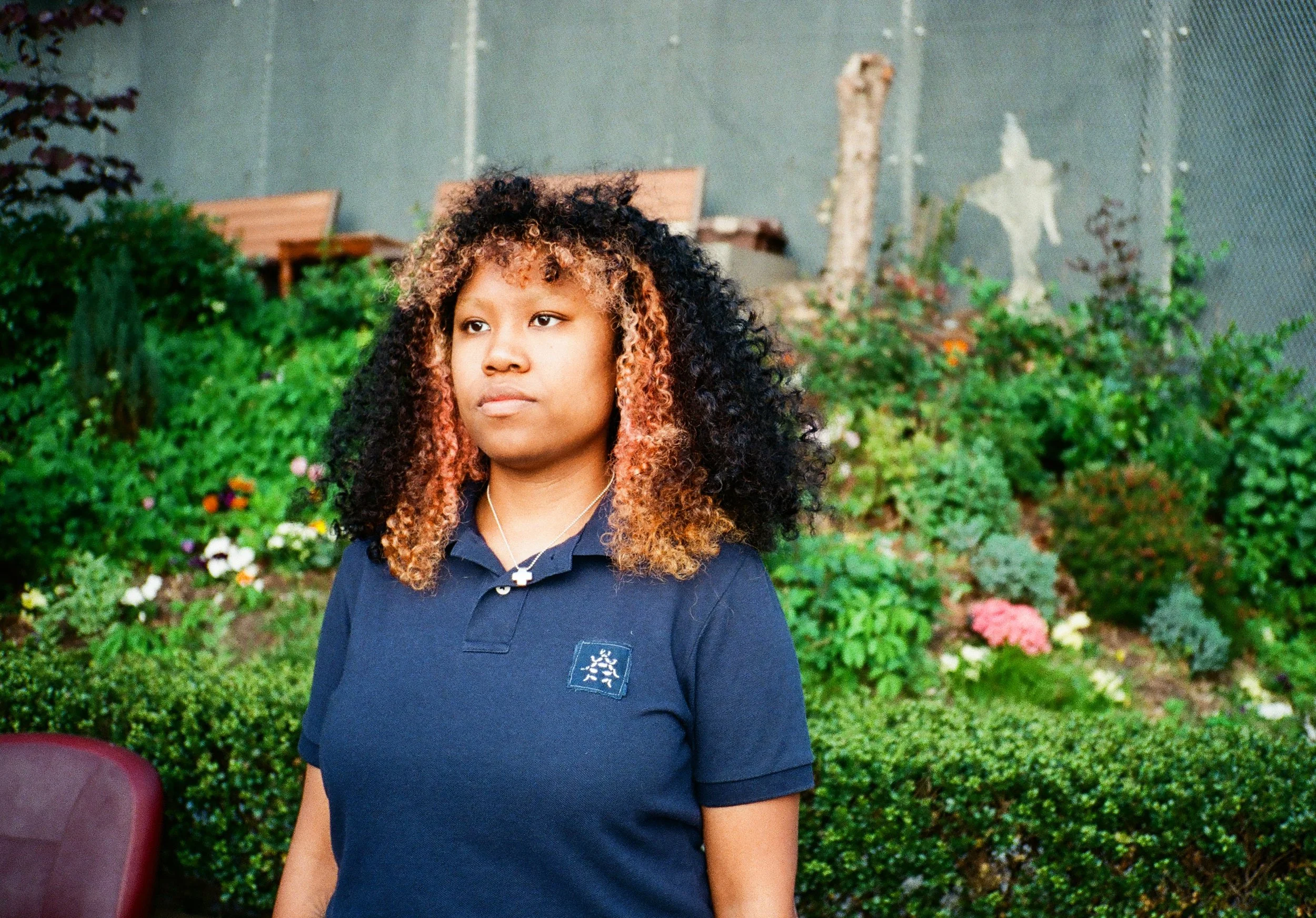 A young woman with curly hair standing outdoors in front of green bushes and colorful flowers, wearing a navy blue polo shirt and a necklace with a cross pendant.