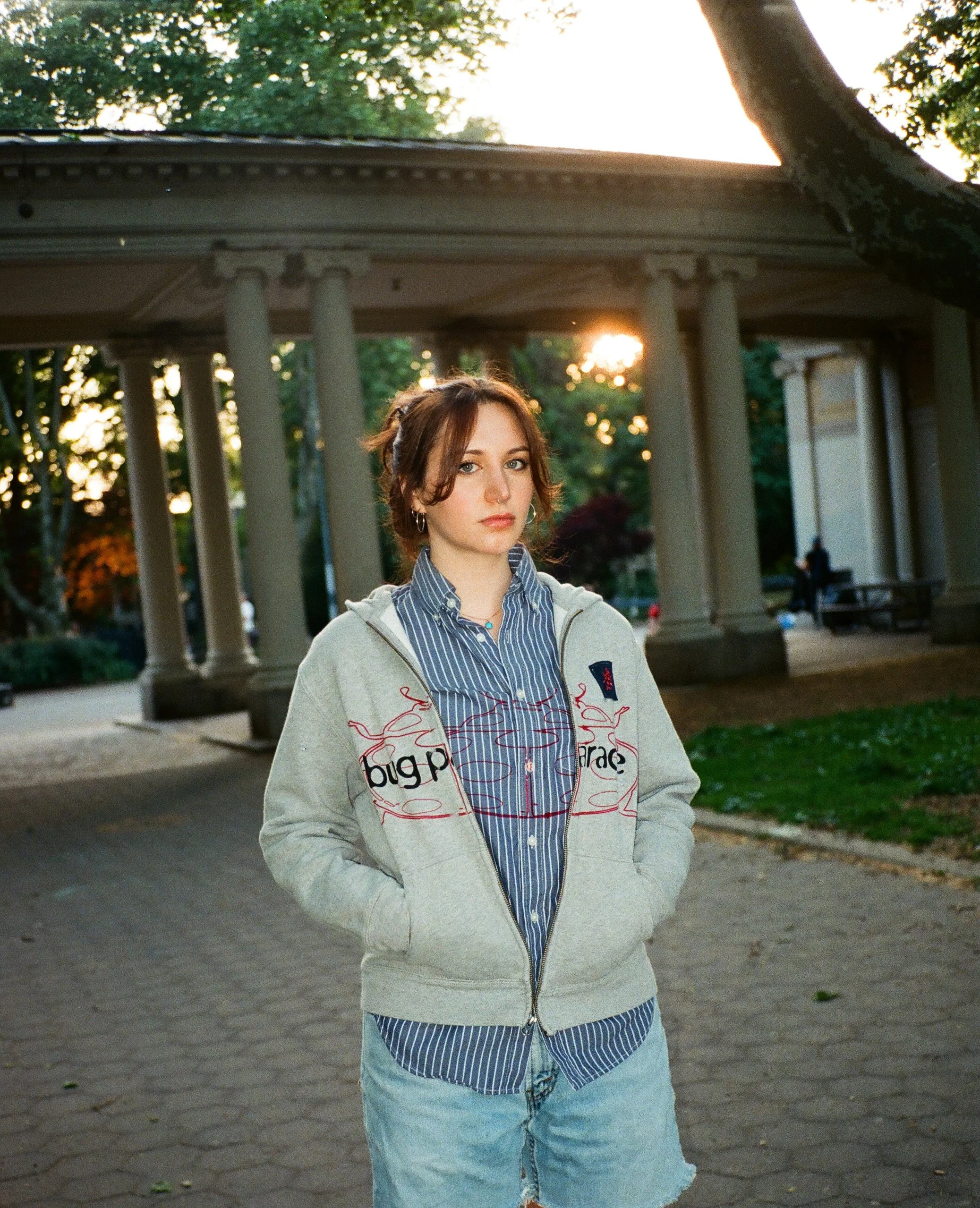 A young woman with brown hair and hoop earrings standing outdoors during sunset, wearing a hoodie with a graphic design and a striped shirt underneath, with a pavilion and trees in the background.