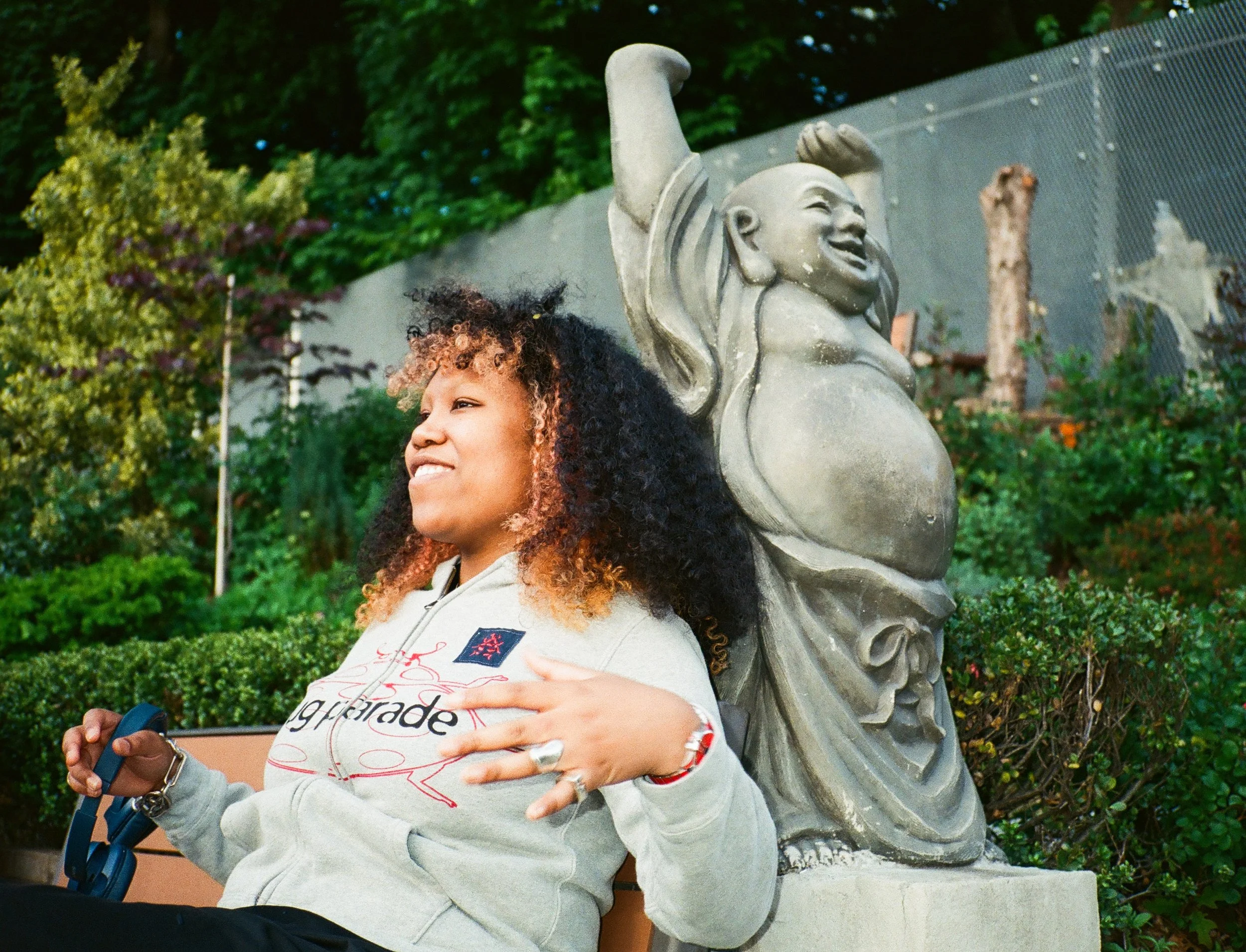 A young woman with curly hair smiling and gesturing with her hands stands near a gray statue of a mermaid in an outdoor park with green trees and bushes in the background.
