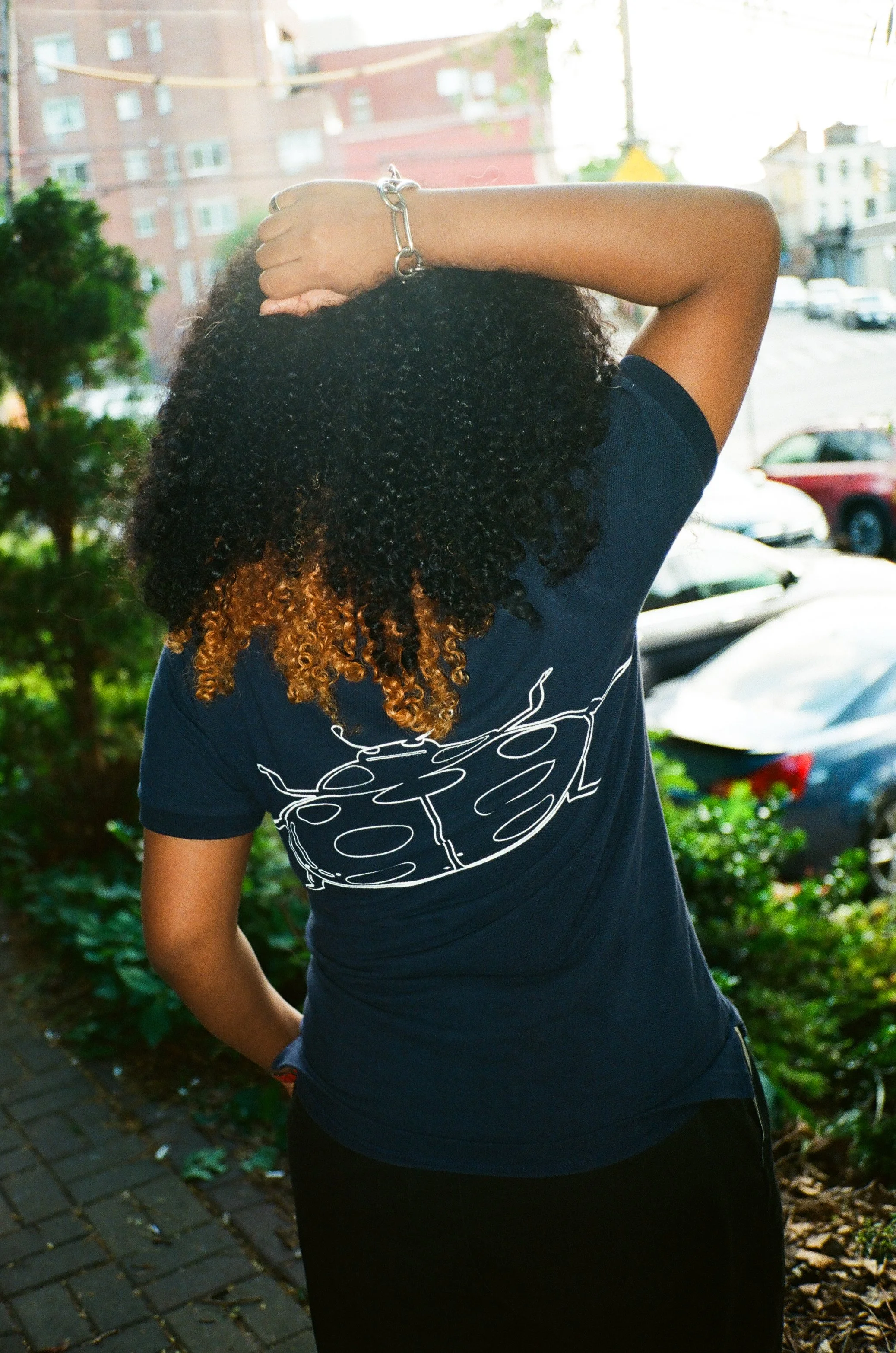 A person with curly black and blonde hair standing outdoors on a brick sidewalk, wearing a black T-shirt with a white graphic design on the back, with one arm raised and resting on their head, jewelry on their wrist, neighborhood buildings and cars i