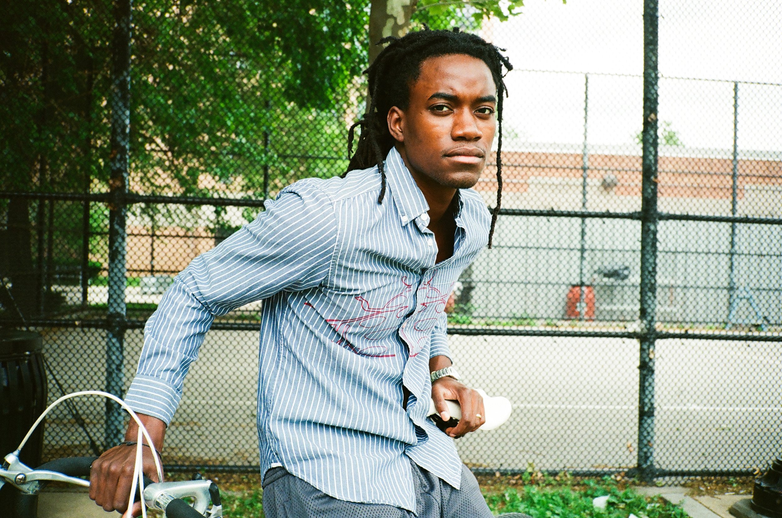 A young man with dreadlocks wearing a light blue striped shirt standing outdoors near a bicycle, with a chain-link fence and trees in the background.