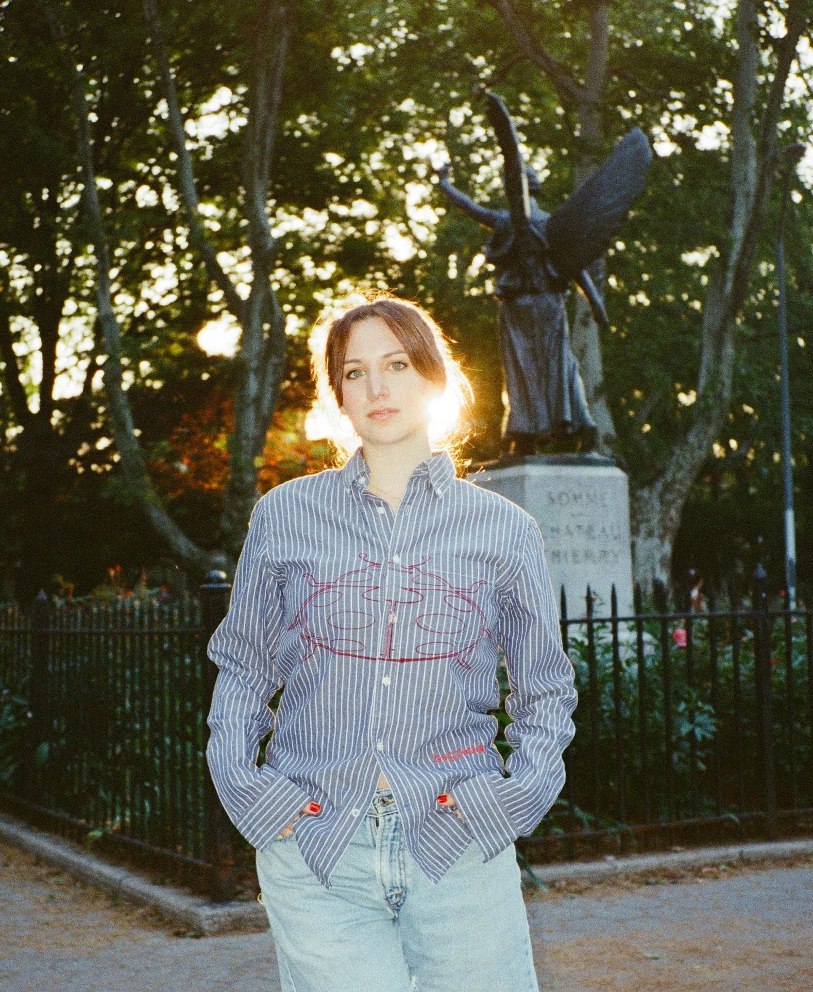 A woman with reddish-brown hair and fair skin is standing outdoors at sunset or sunrise, with trees and a statue in the background. She is wearing a blue and white striped shirt with embroidery, and light blue jeans. Her hands are in her pockets, and