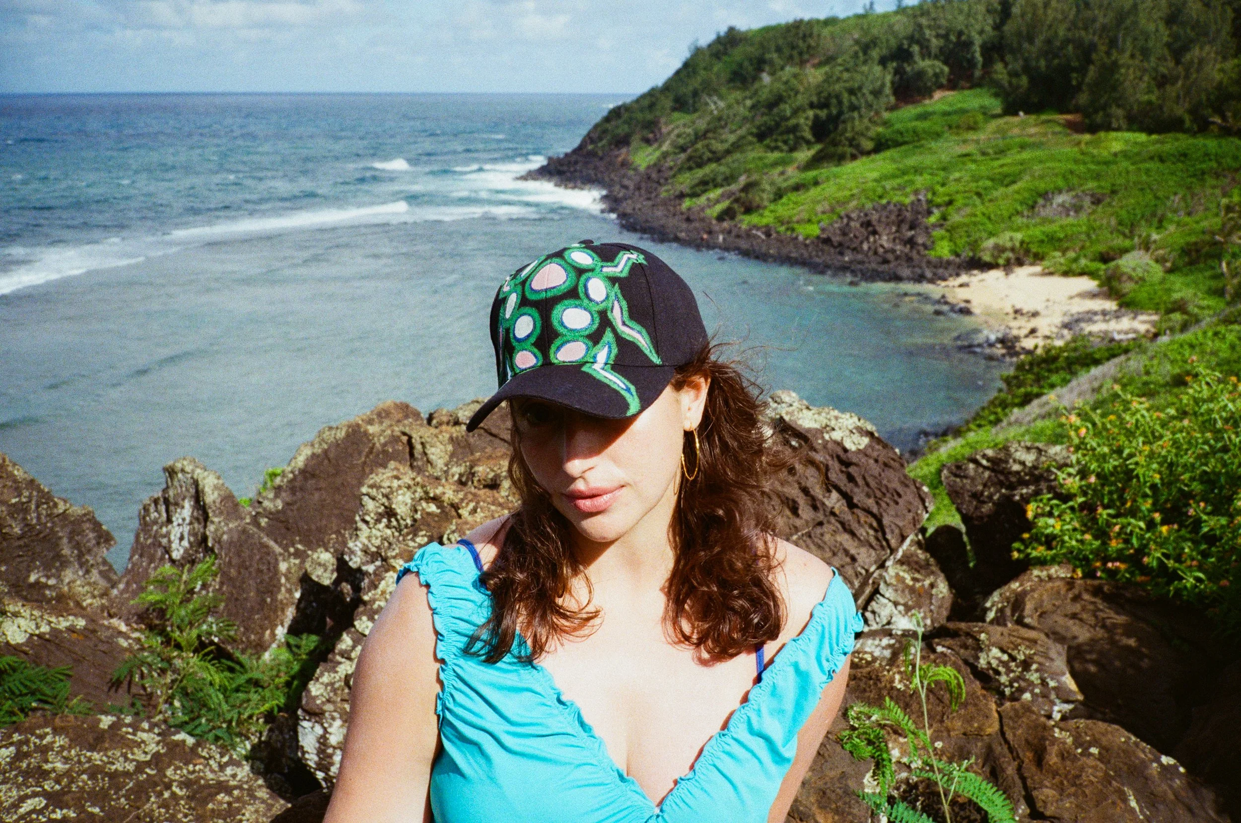 A woman with curly brown hair wearing a blue off-the-shoulder top and a colorful cap with green, white, and pink patterns, standing in front of a coastal cliff with rocks, green vegetation, and the ocean in the background.