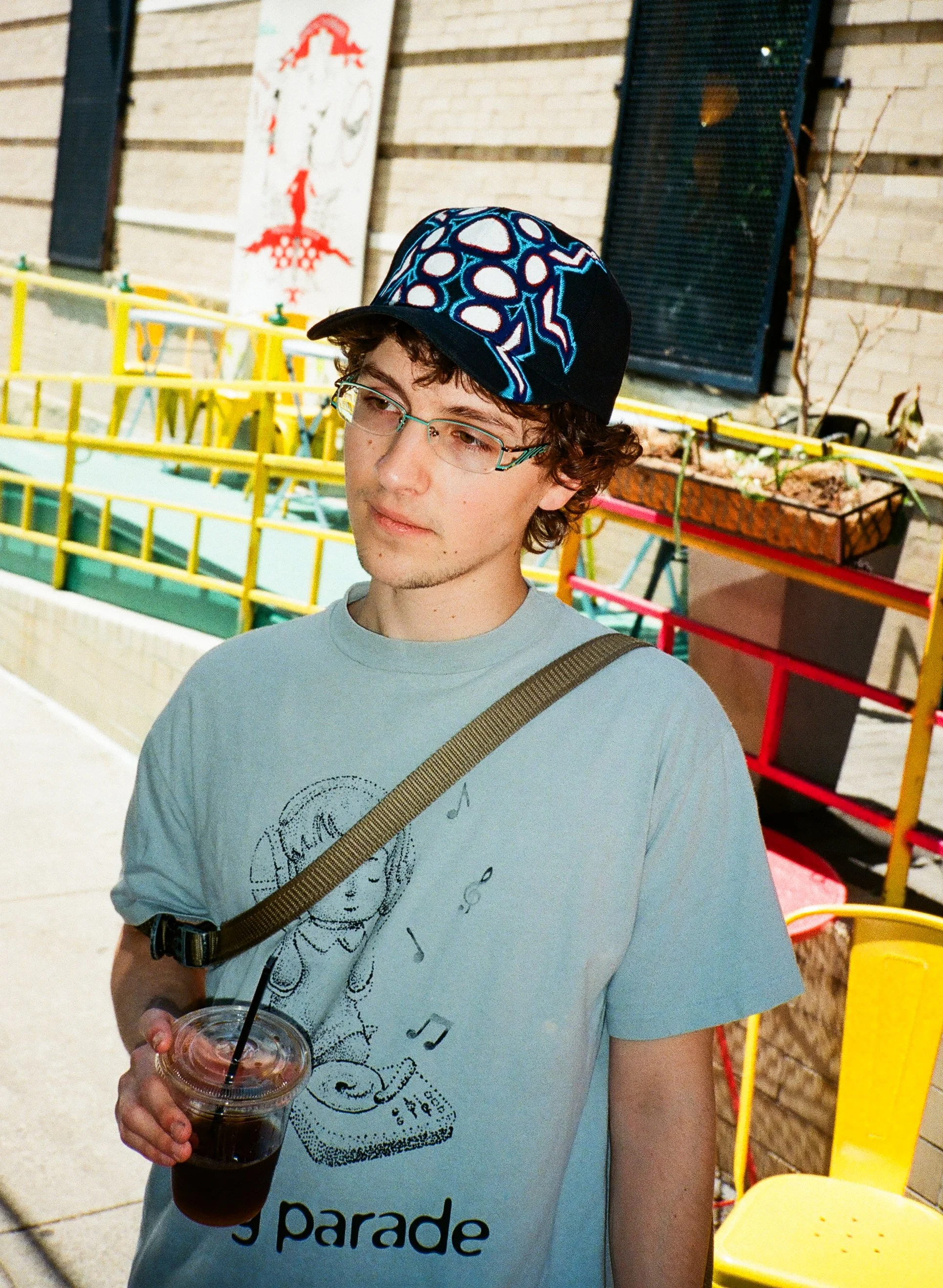 A young person with curly hair, glasses, and a graphic T-shirt holding a plastic cup of dark-colored soda with a straw in what appears to be an indoor or semi-indoor setting, with colorful railings and a brick wall in the background.