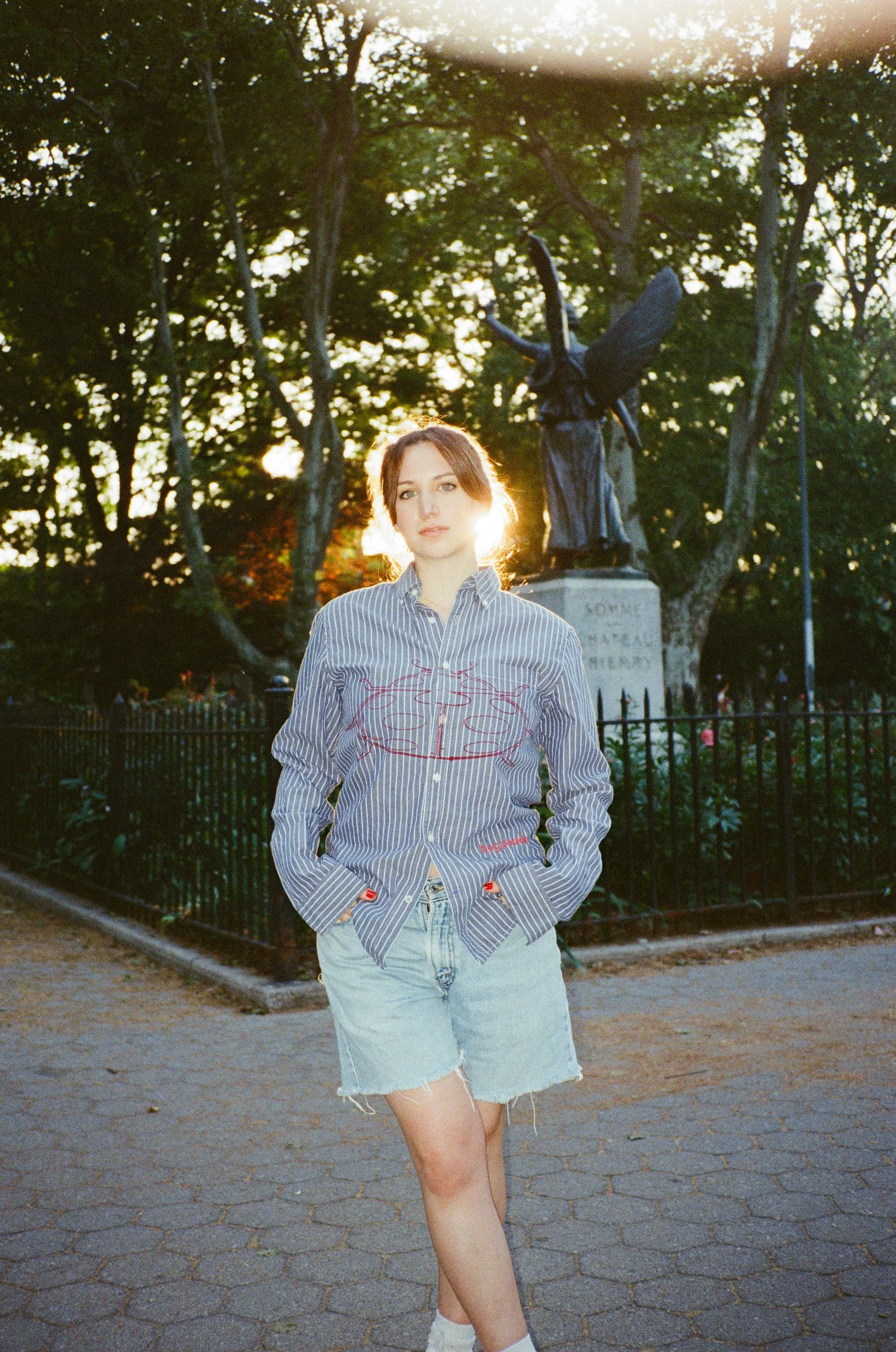 A young woman with red hair wearing a striped shirt and ripped denim shorts standing on a paved path in a park during sunset. In the background, there is a statue of a figure with wings, trees, and a black fence.