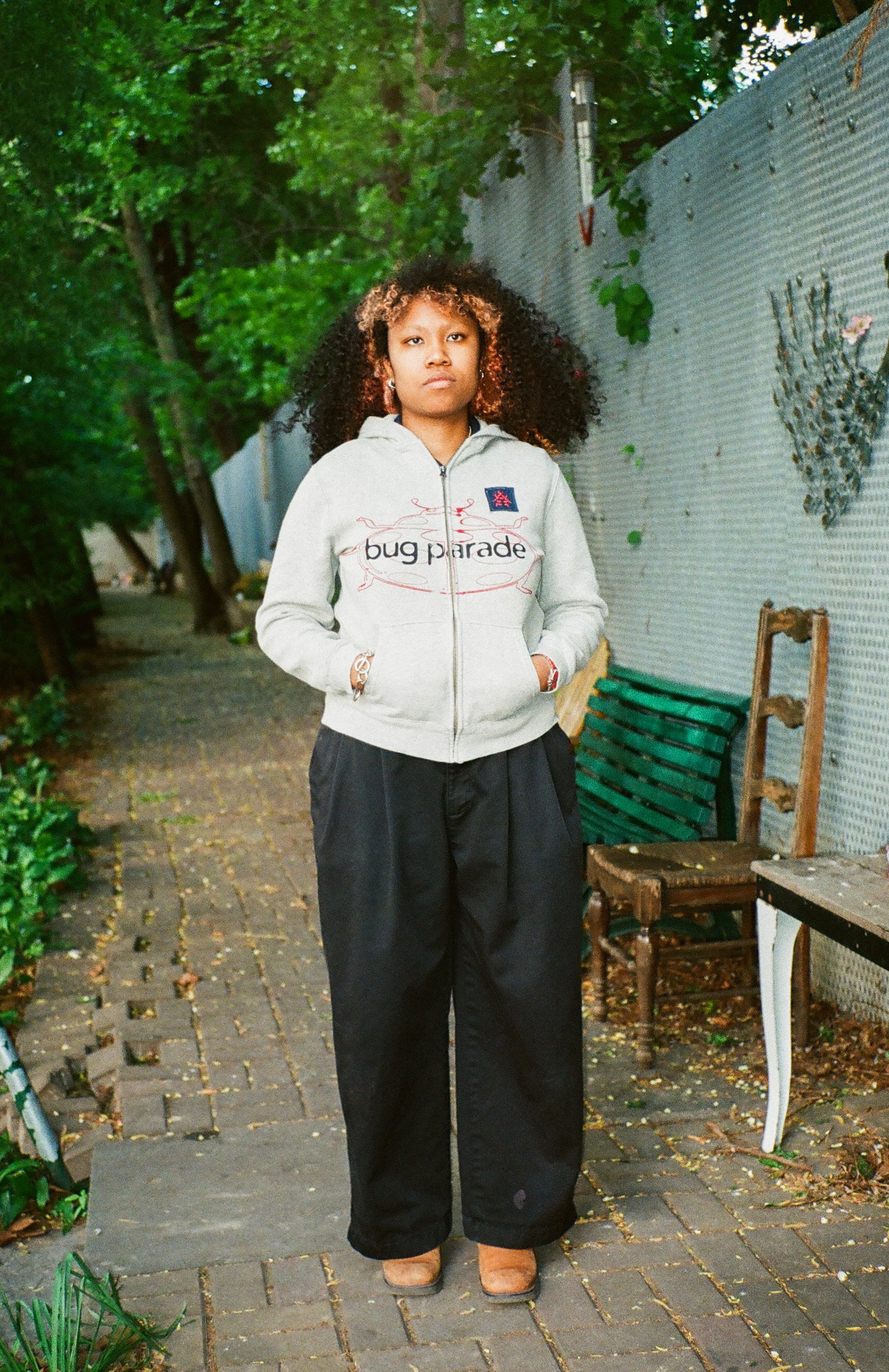 A woman standing on a brick path outdoors, wearing a light gray hoodie with 'bug parade' written on it, black wide-leg pants, and brown boots. She has curly hair with blonde highlights and a serious expression, with green fencing and trees in the bac