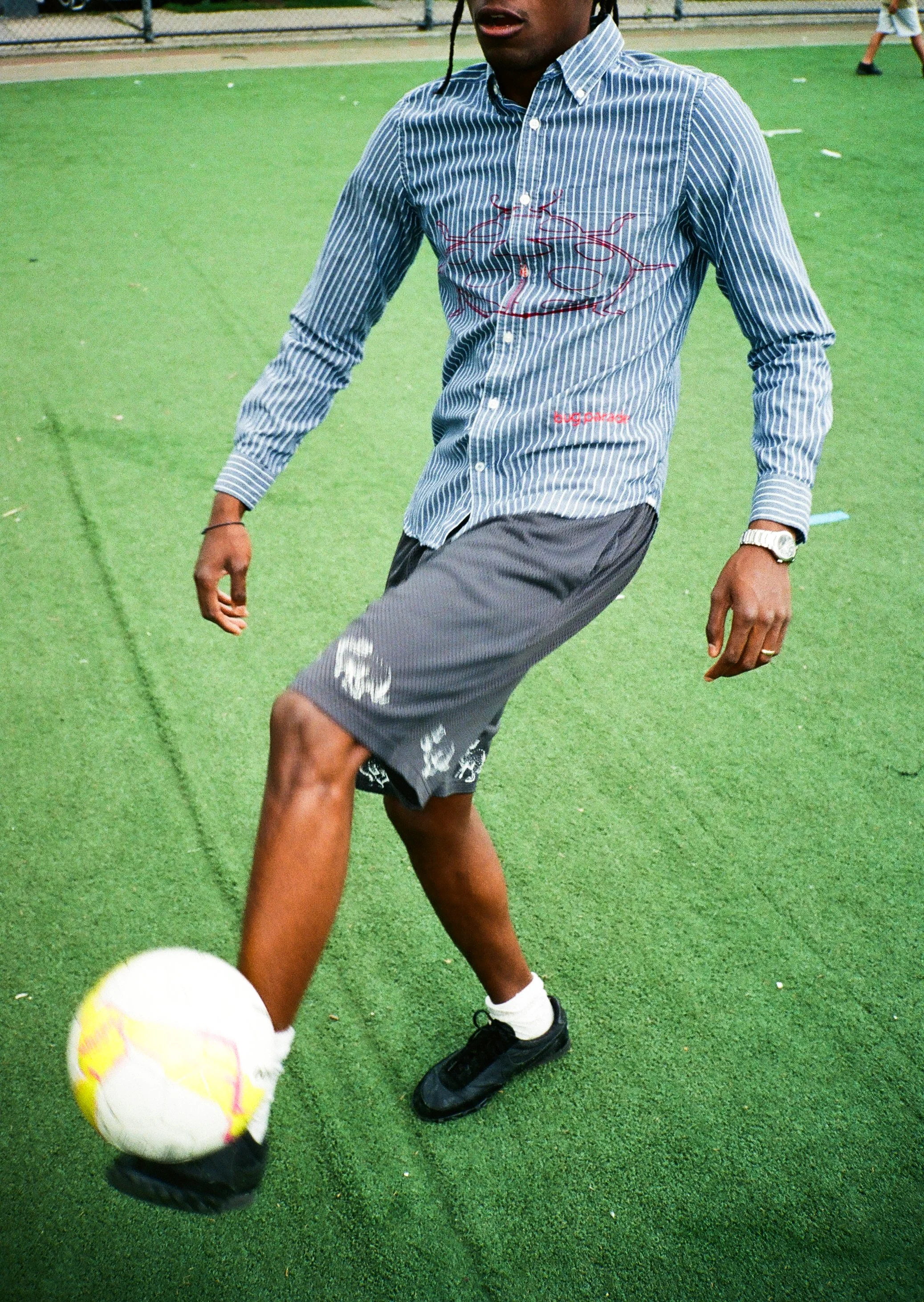 A person wearing a striped long sleeve shirt and gray shorts is playing soccer on a green artificial turf field. Someone's arm and hand are visible in the foreground, reaching towards a soccer ball.