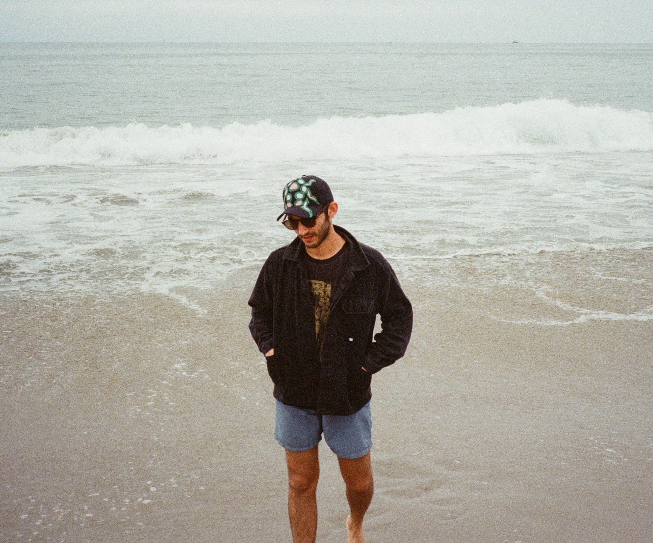 A man walking on the beach, wearing a black jacket, shorts, sunglasses, and a baseball cap with a green and white polka dot pattern. The ocean with gentle waves is in the background.