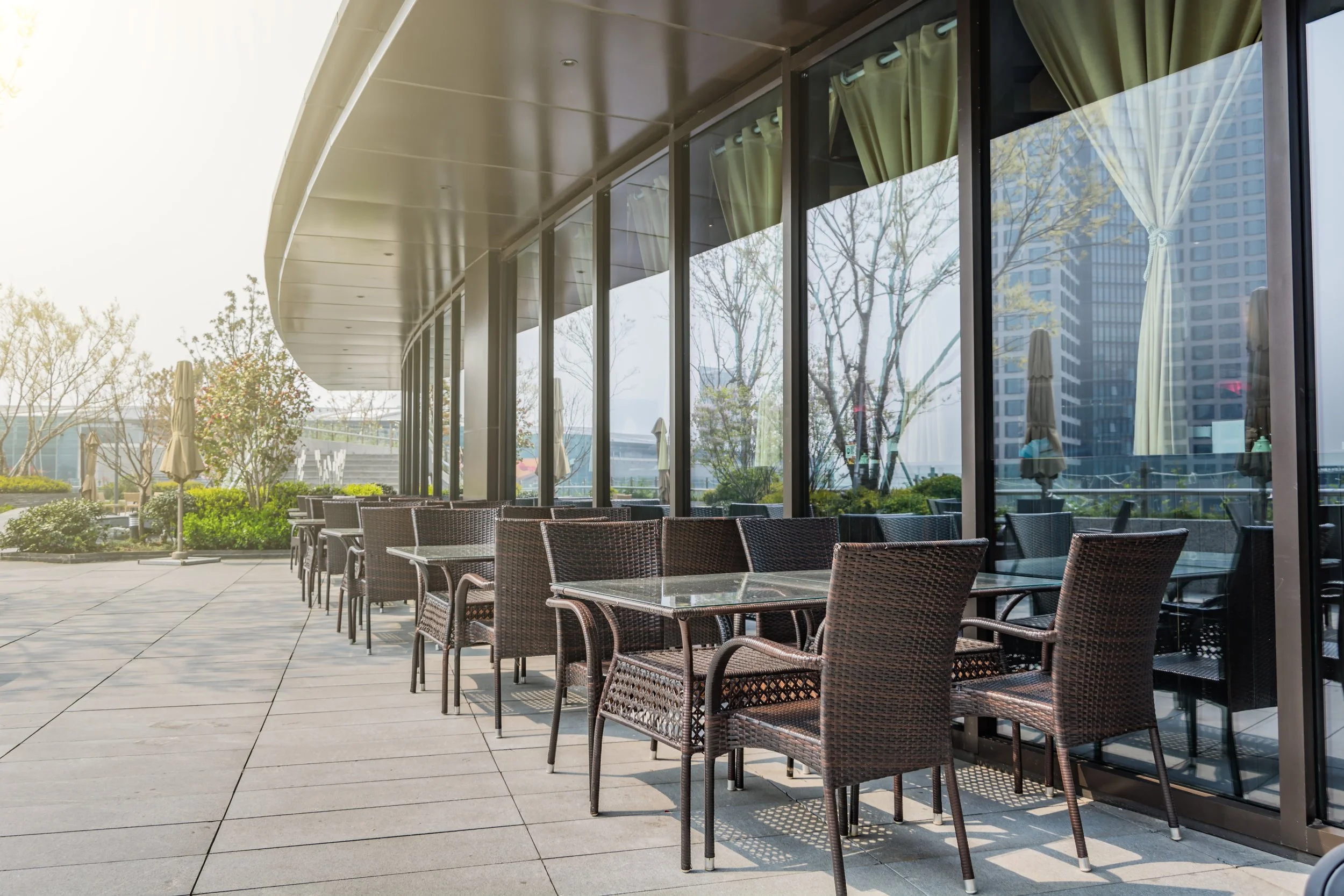 Terrasse d'un restaurant avec des tables en verre et des chaises en rotin, vue sur des bâtiments modernes et des arbres, intérieur visible à travers les grandes fenêtres avec des rideaux verts.