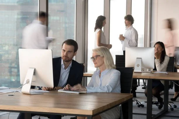 Deux personnes travaillant sur un ordinateur dans un bureau moderne avec des fenêtres panoramiques, d'autres collègues discutant en arrière-plan.