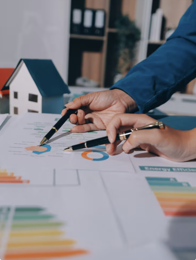 Deux personnes discutent autour de documents financiers et graphiques, avec une petite maison en décoration en arrière-plan.