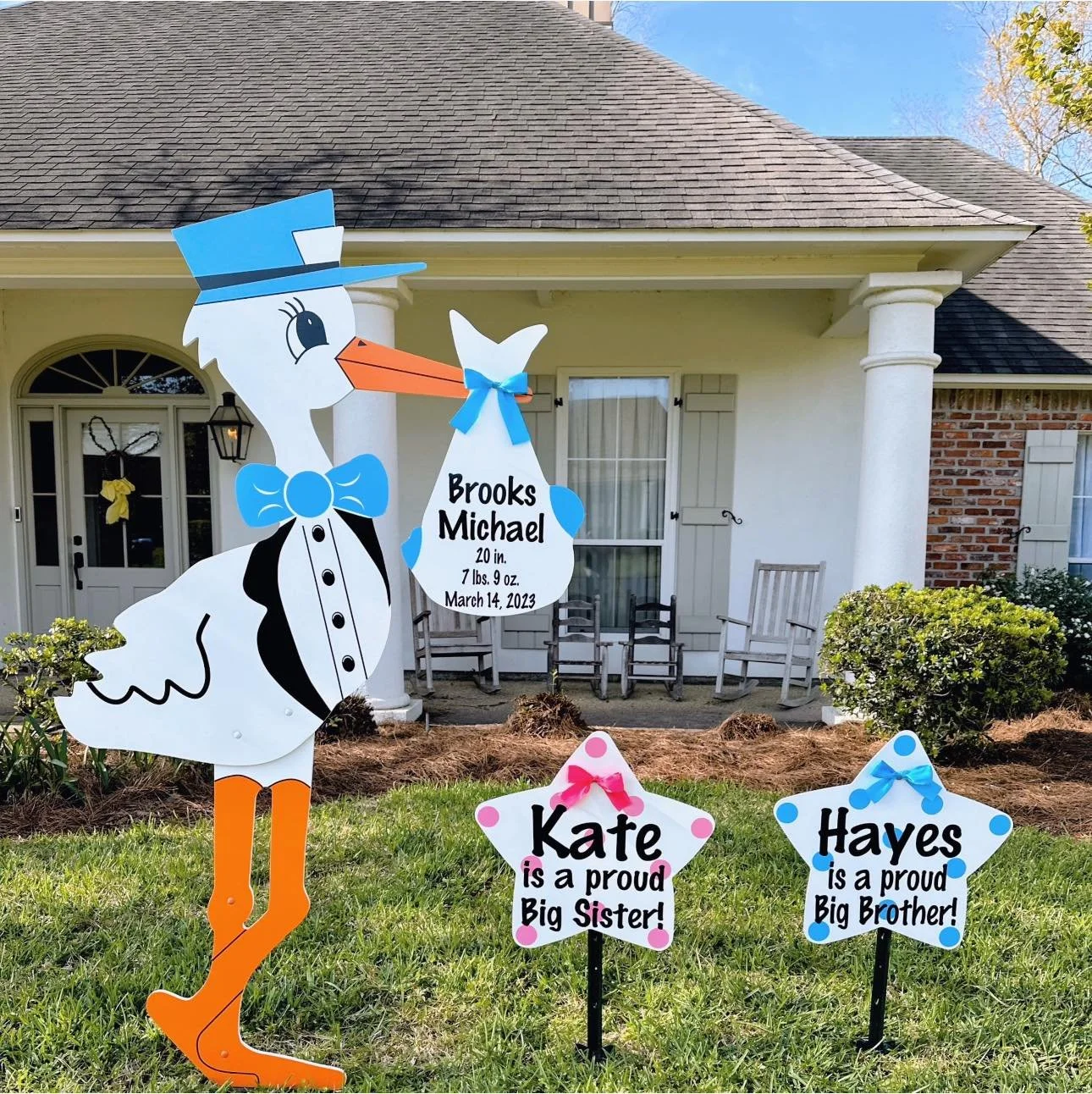 Decorative outdoor display with a large cartoon-style stork holding a sign with newborn baby details, and two star-shaped signs honoring siblings, set in front of a house with bushes and chairs on the porch.
