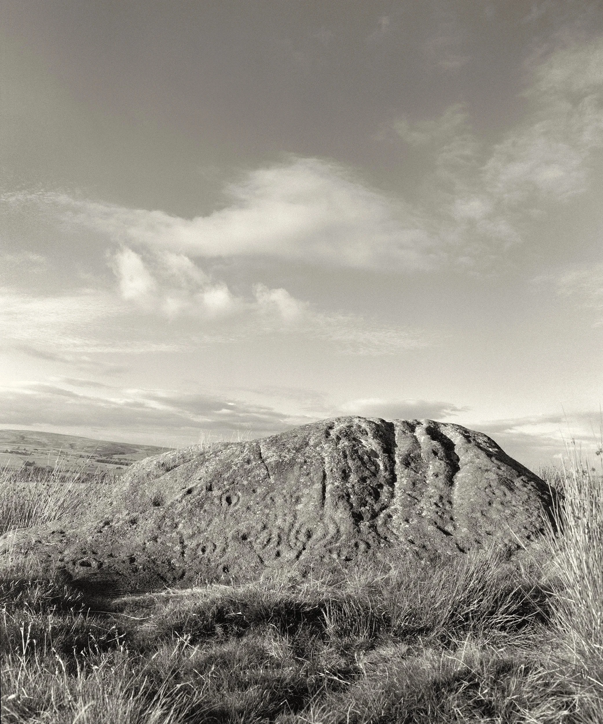 A black and white photographic image of a rock shaped like a giant badger with prehistoric cup and ring carvings, photographed against an atmospheric Yorkshire sky | a photograph entitled Badger Stone, situated on Ilkley Moor, Yorkshire.