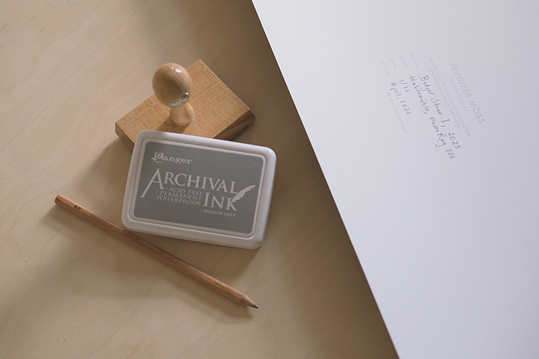 Photograph of a wooden stamp and archival ink pad with a wooden pencil next to the back of a print showing a filled in Reindeer Moss Photography stamp