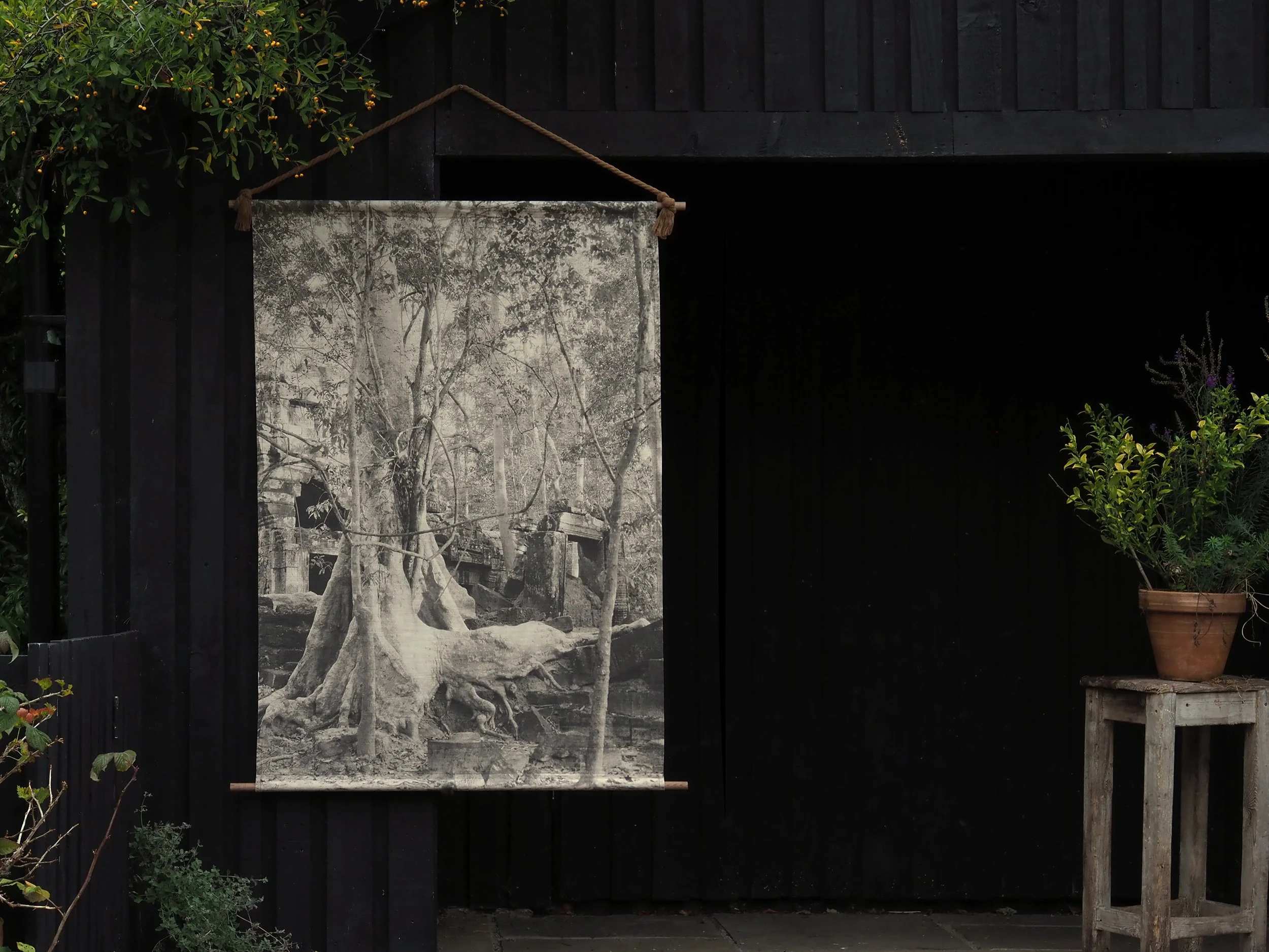 Photographic image of a large scale linen wall hanging against charcoal wood, with natural caramel coloured rope and wood poles, featuring vintage style image of large tree with roots growing over ancient temple ruins of Ta Nei, Angkor in Cambodia