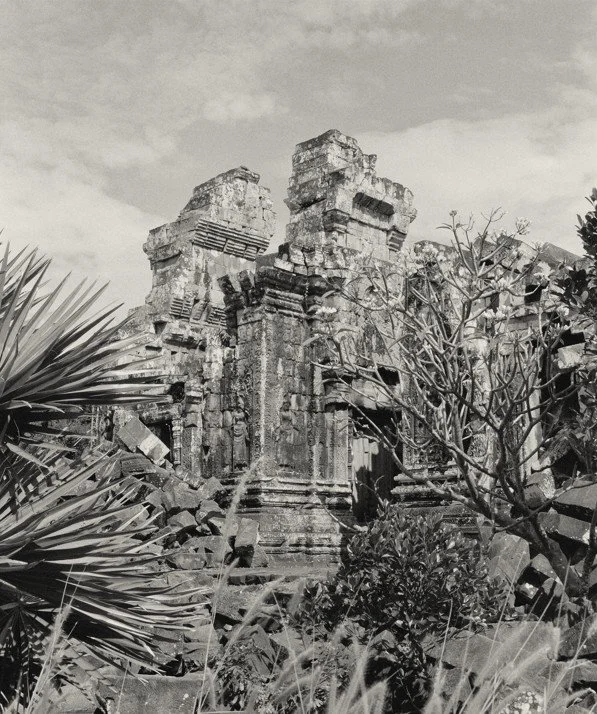 Black and white photographic image of majestic ancient ruins from behind tropical foliage in the ancient Angkor site of Phnom Bok in Cambodia